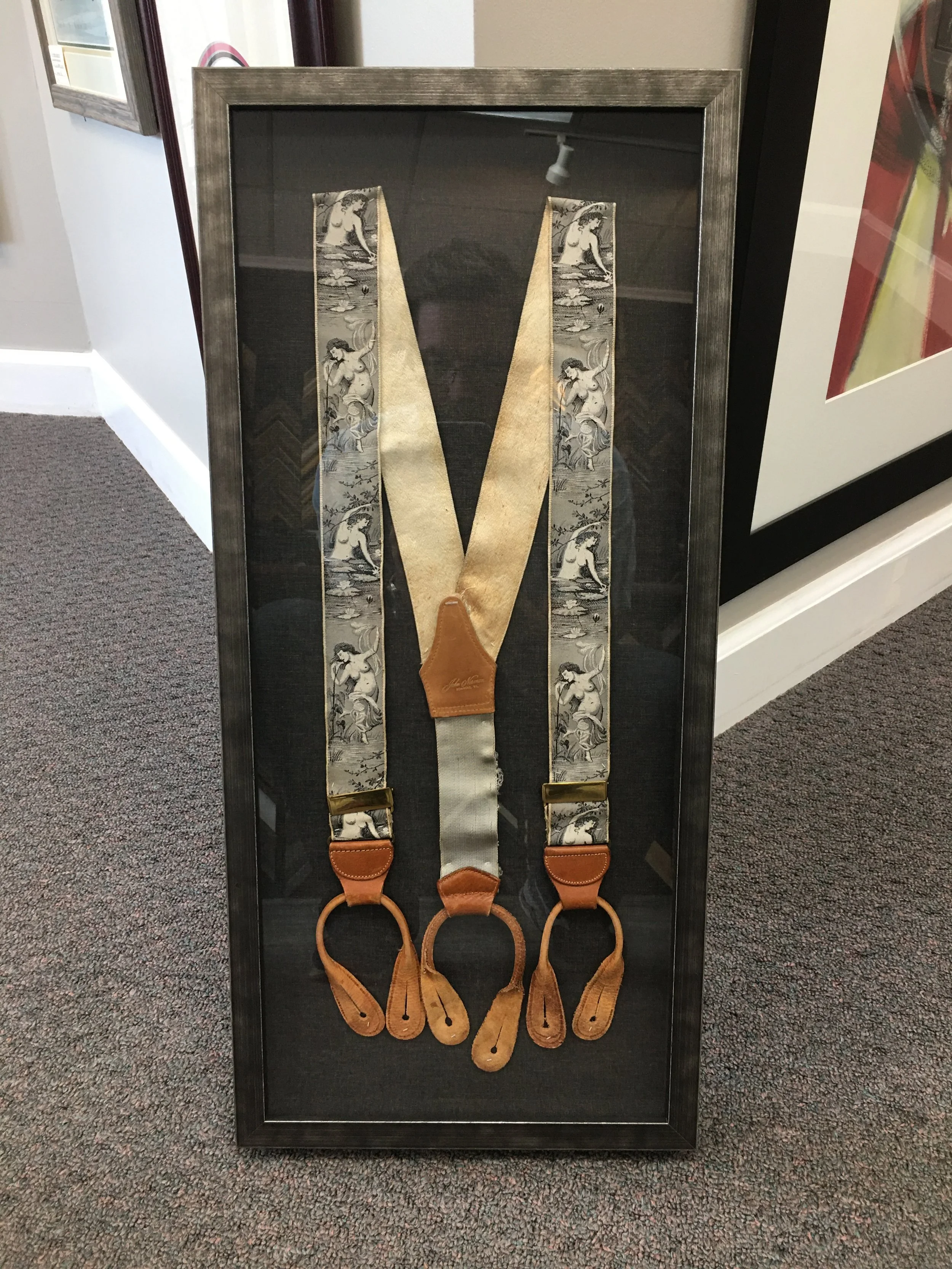 Display of decorative shoelace suspenders with a vintage pattern and leather accents in a frame.