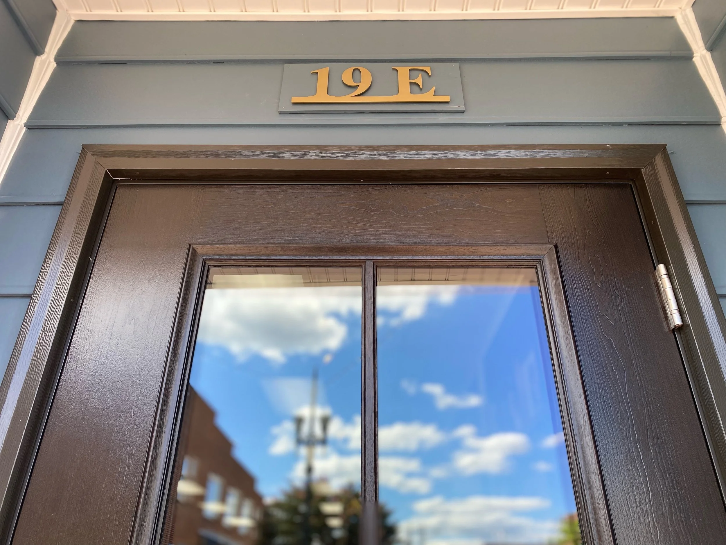 Close-up of a door with a saint glass panel reflection, titled '19 E' sign above the door, blue wall, outdoor scene with blue sky and clouds.