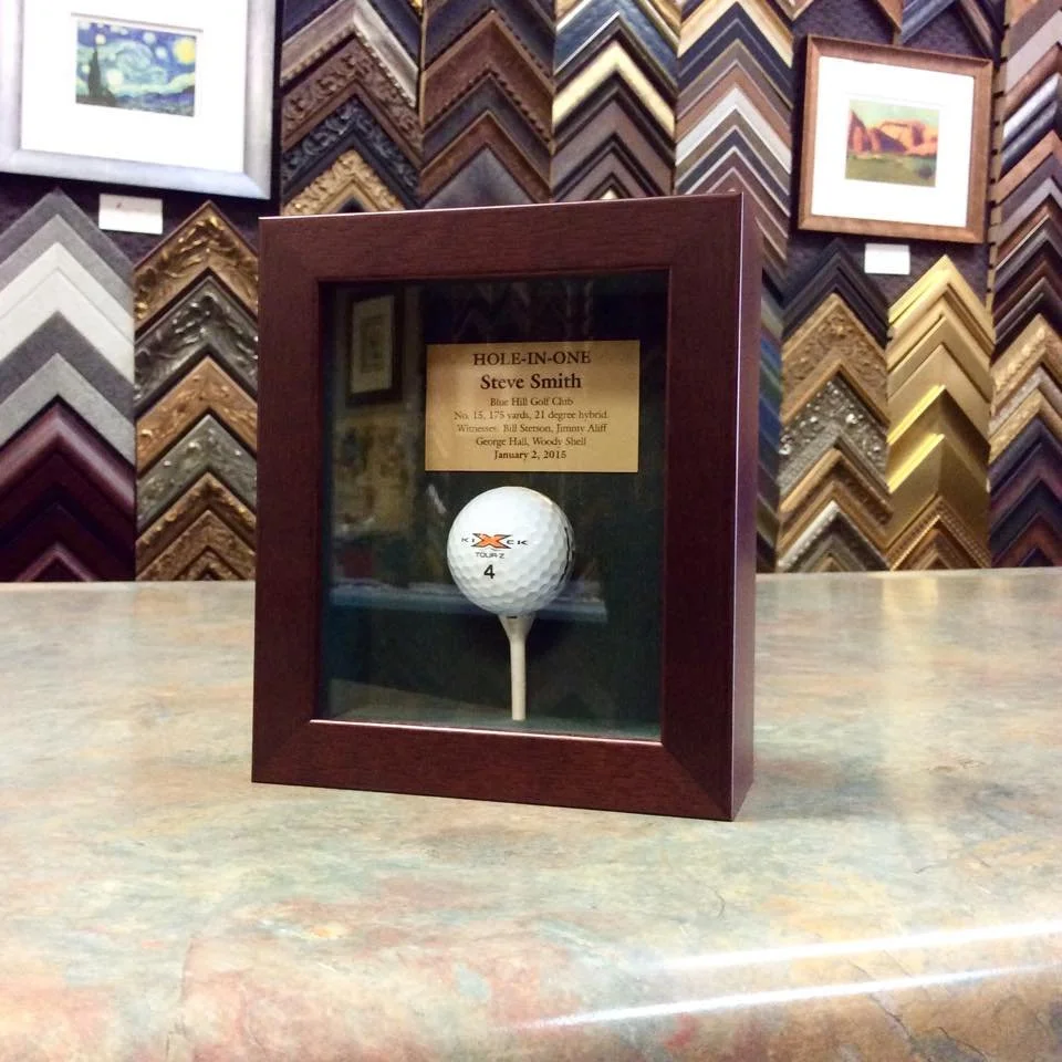 A framed display with a golf ball and a plaque inside, set on a wooden surface in front of a wall decorated with many picture frame samples.