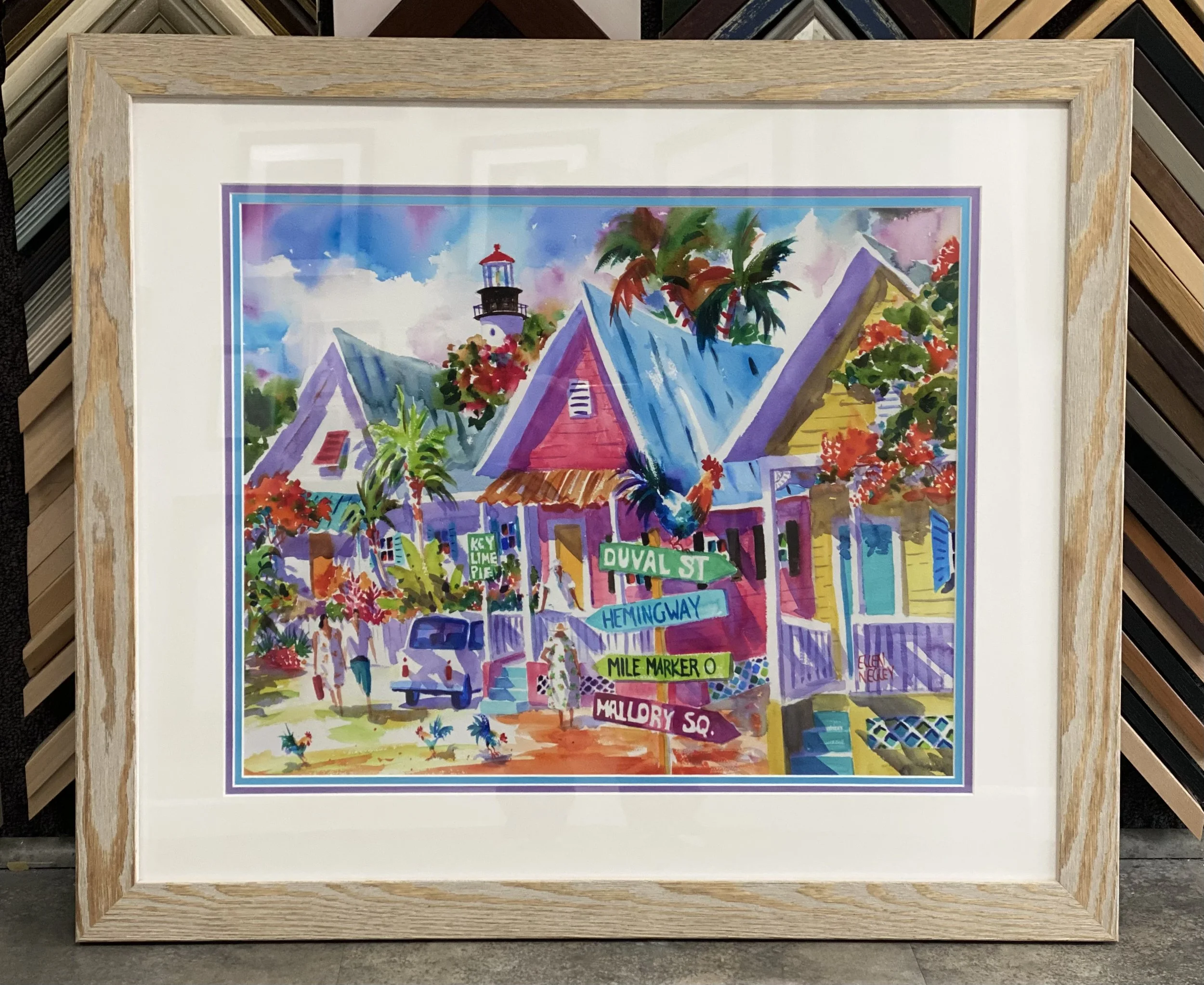 Colorful watercolor painting of a neighborhood scene with houses, palm trees, street signs, and a lighthouse, framed with a wooden frame, displayed in a store or gallery.