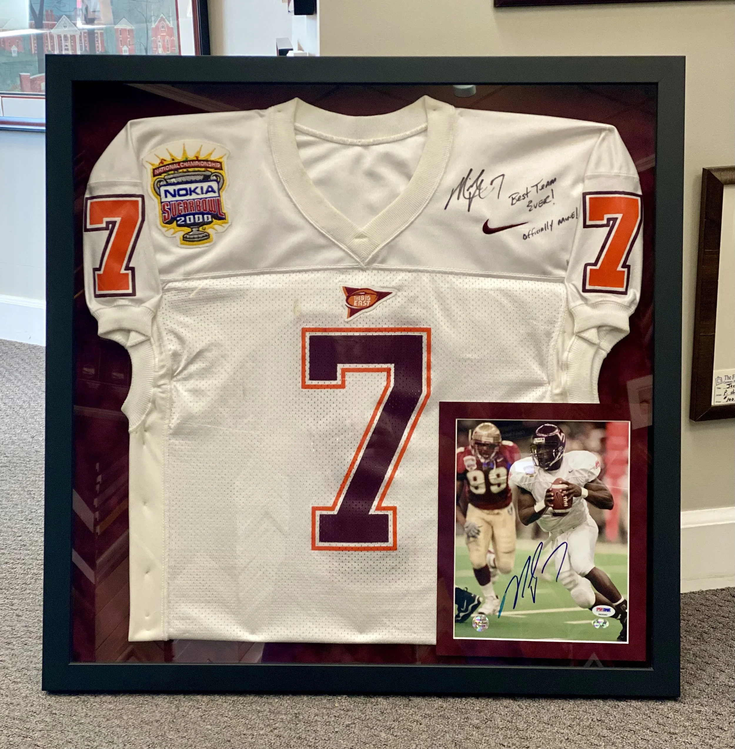 A framed display of a white football jersey signed by a player, featuring patches and number 21, with a photograph of a football game at the bottom right corner.