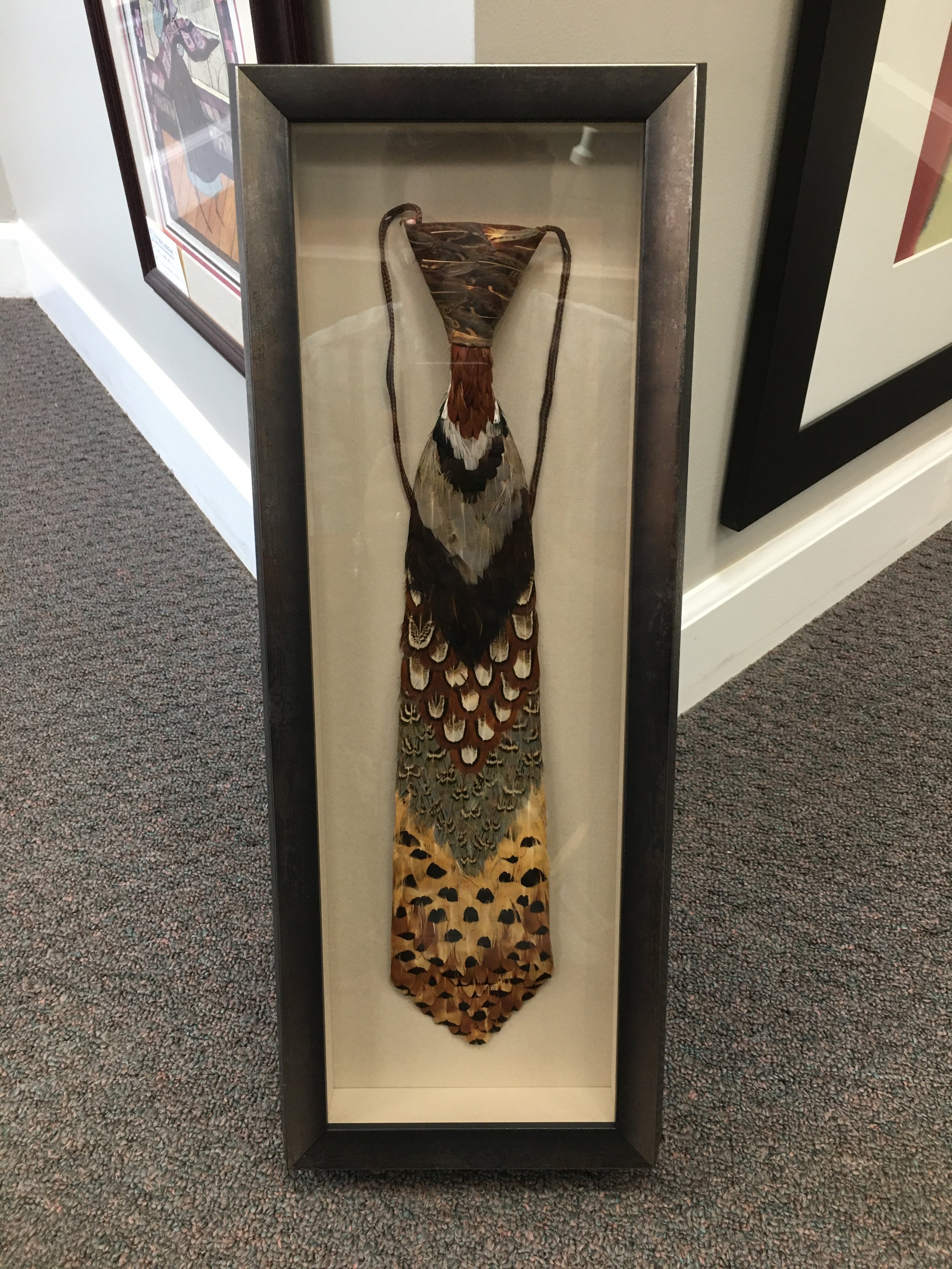 A framed display of a peacock feather headdress with a multicolored, patterned arrangement of feathers, mounted on a white background.