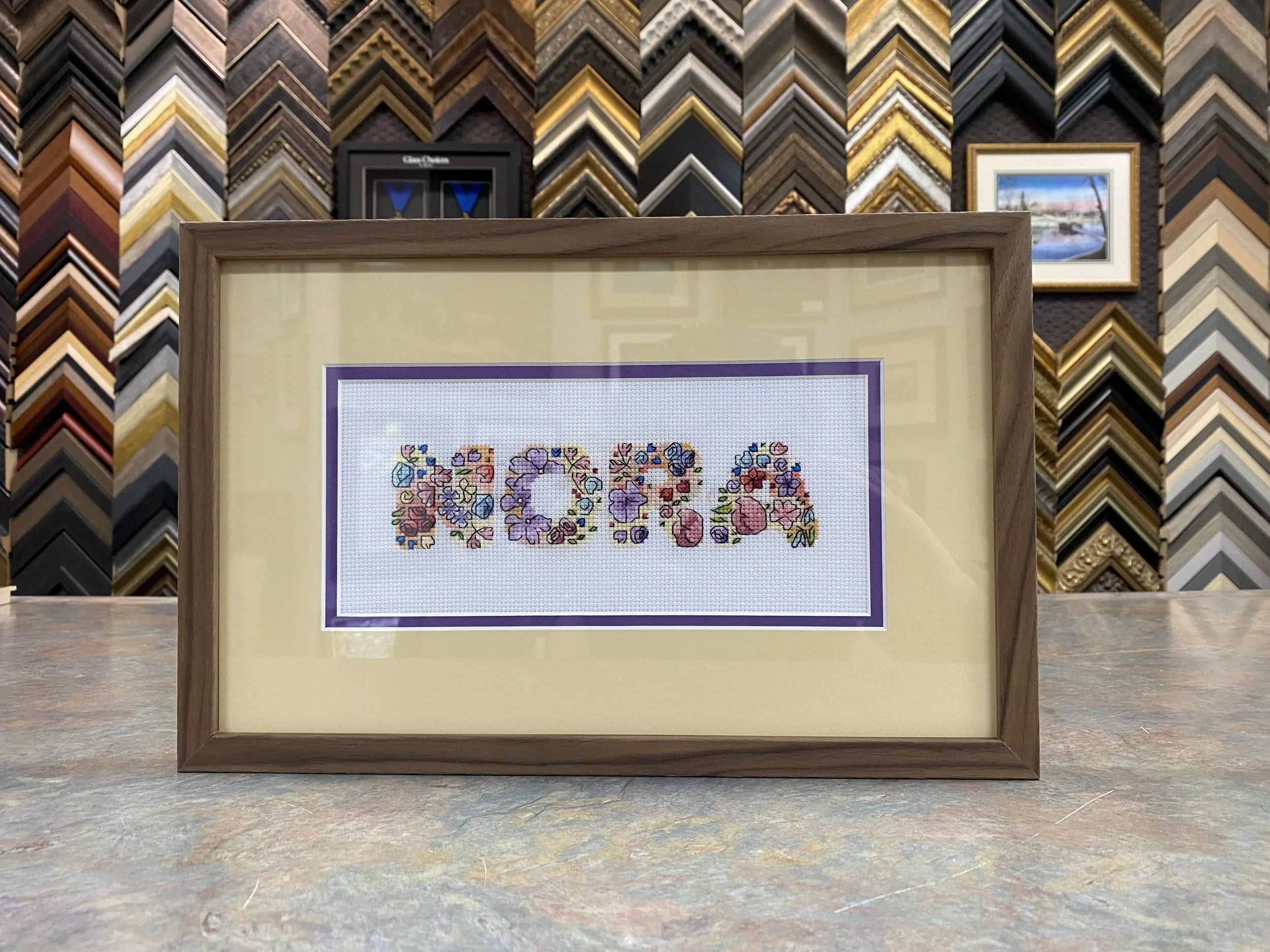 Framed cross-stitch embroidery of the word "LOVE" made up of colorful flowers, displayed on a yellow background.