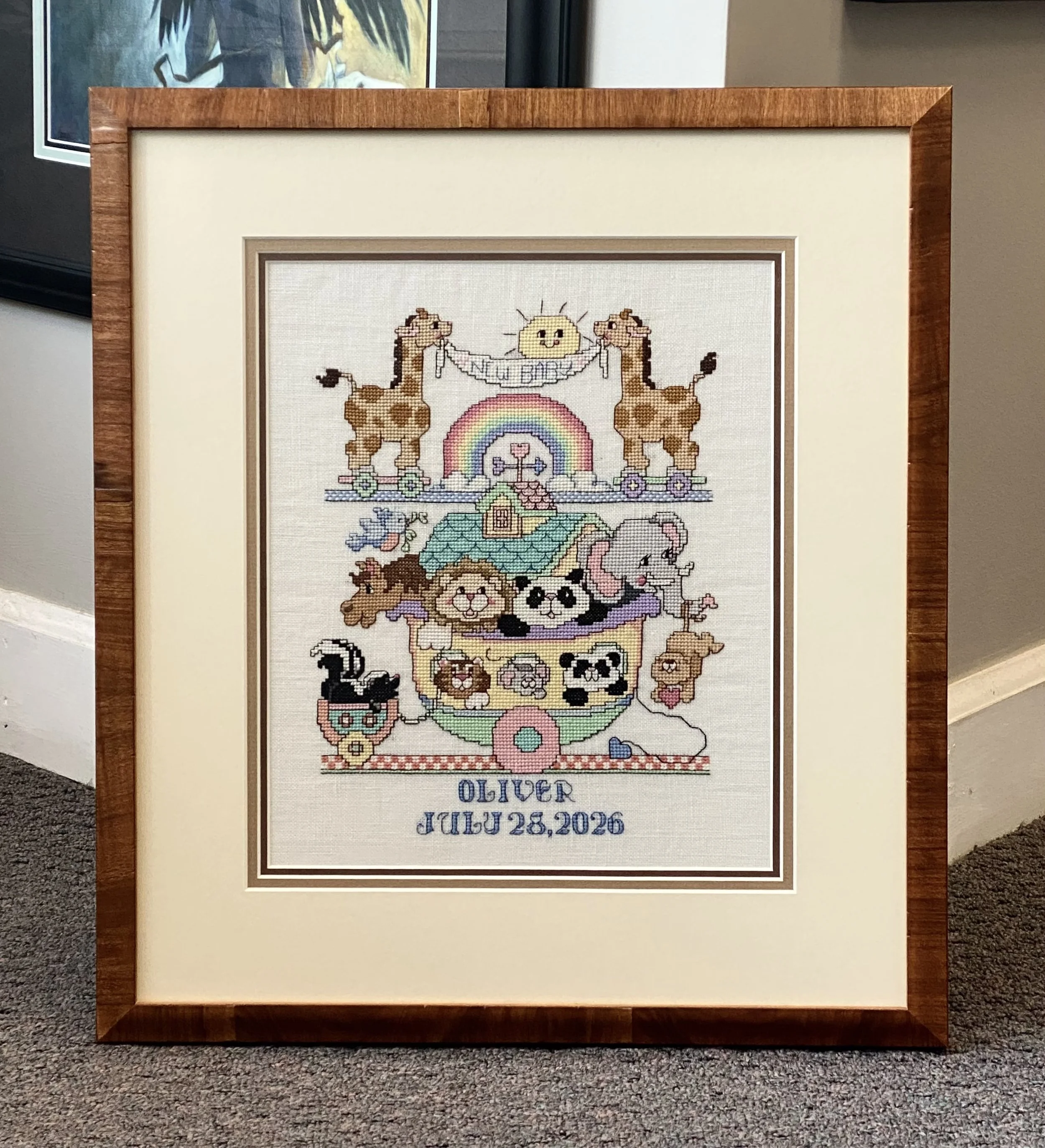 Framed cross-stitch of Noah's Ark with animals celebrating the birth of a child using a cream mat and medium brown olive wood frame.
