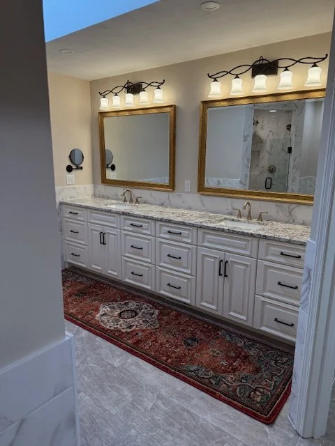 Double bathroom vanity with white cabinets, granite countertop, two mirrors with gold frames, and four light fixtures above each mirror in a bathroom. There is a red patterned rug on the floor. Expert custom mirror framing in Salem, VA, and near Roan
