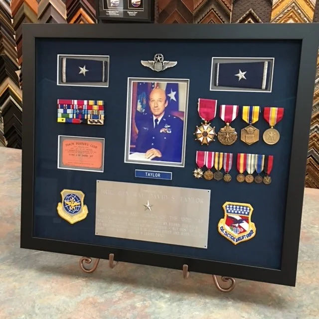 Military shadow box display with a framed photograph of a man in uniform at the center, surrounded by medals, ribbons, patches, and a nameplate labeled 'Taylor.'