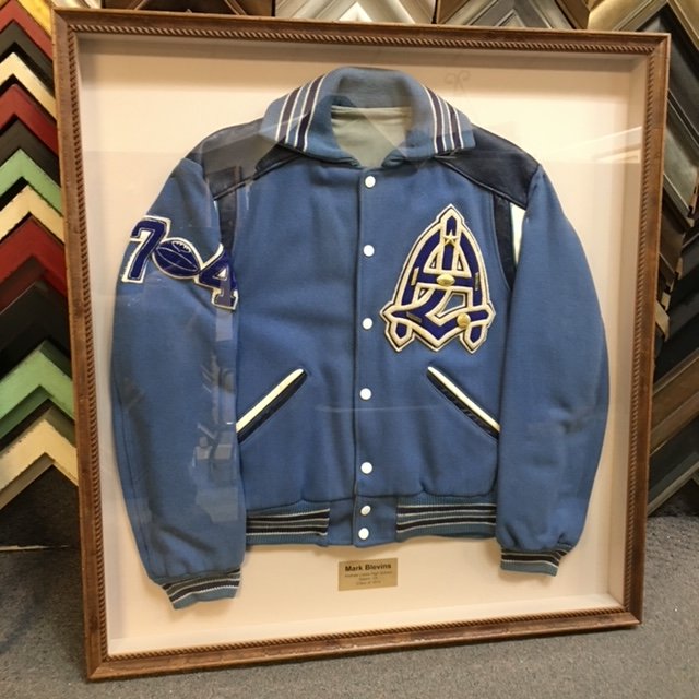 Framed classic blue varsity jacket with white and black accents, featuring a letter A on the chest and the number 74 on the sleeve, displayed in a store.