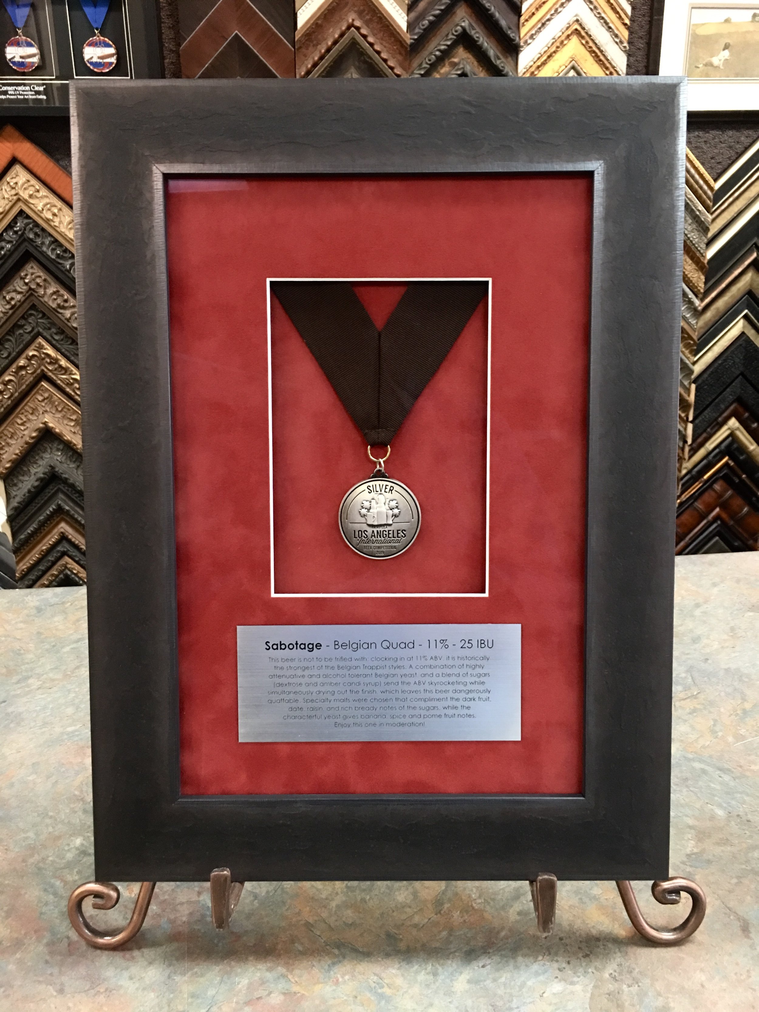 A framed medal display with a black ribbon and a silver medal inscribed with "Silver Los Angeles International." The display is set against a red background with a description placard below. The background features various picture frames and framing samples.