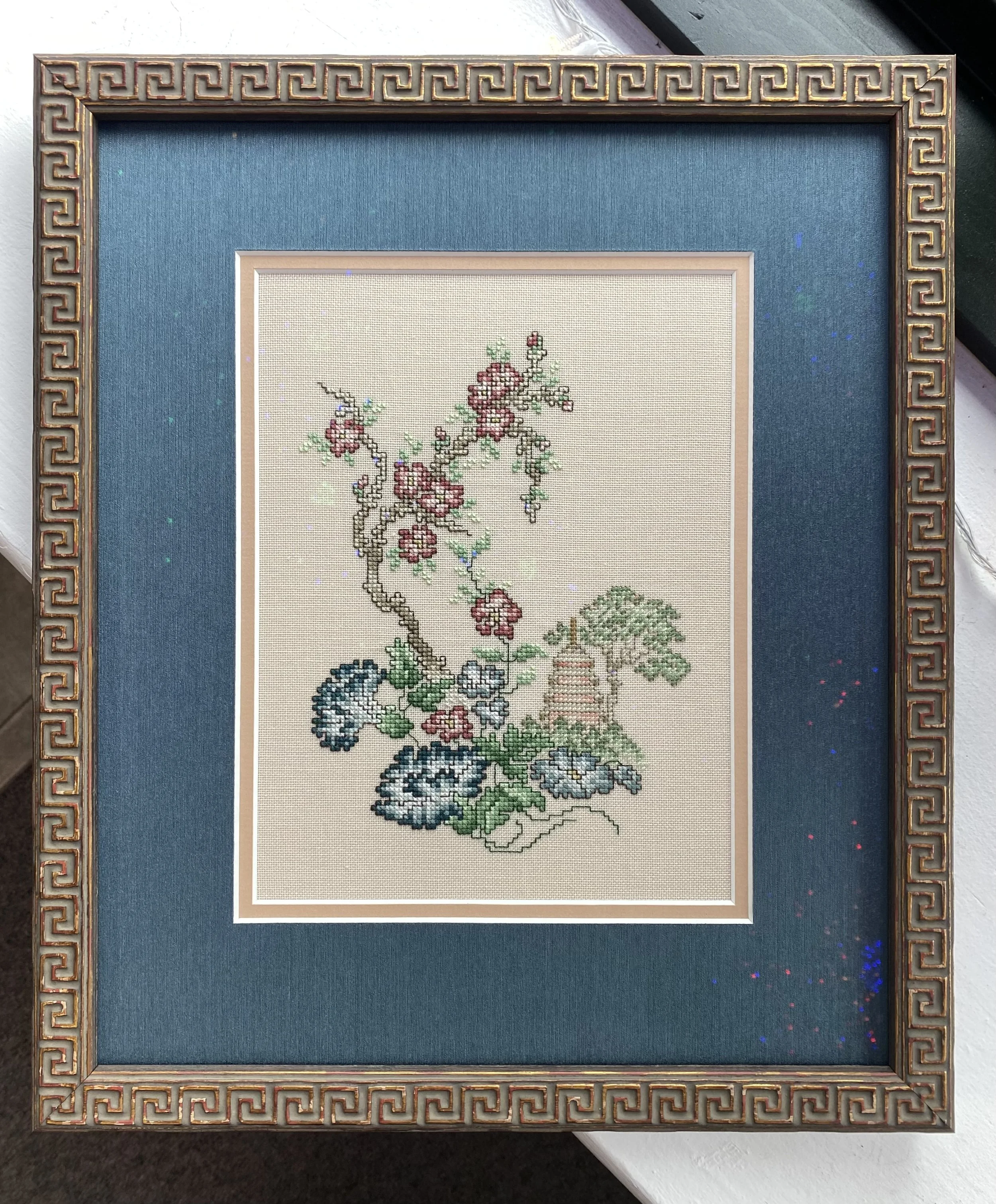 A framed embroidery artwork featuring a blossoming branch with pink flowers and green leaves, set against a cream-colored background, surrounded by a dark blue mat and an ornate wooden Thai-style carved frame.