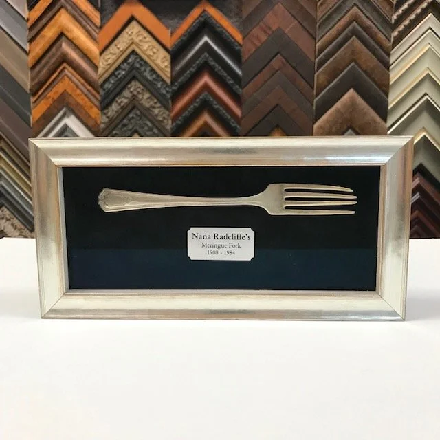 A framed silver fork with a plaque that reads Nana Radcliffe's Menorque Fork 1908 - 1984, displayed against a background of various picture frame moldings.