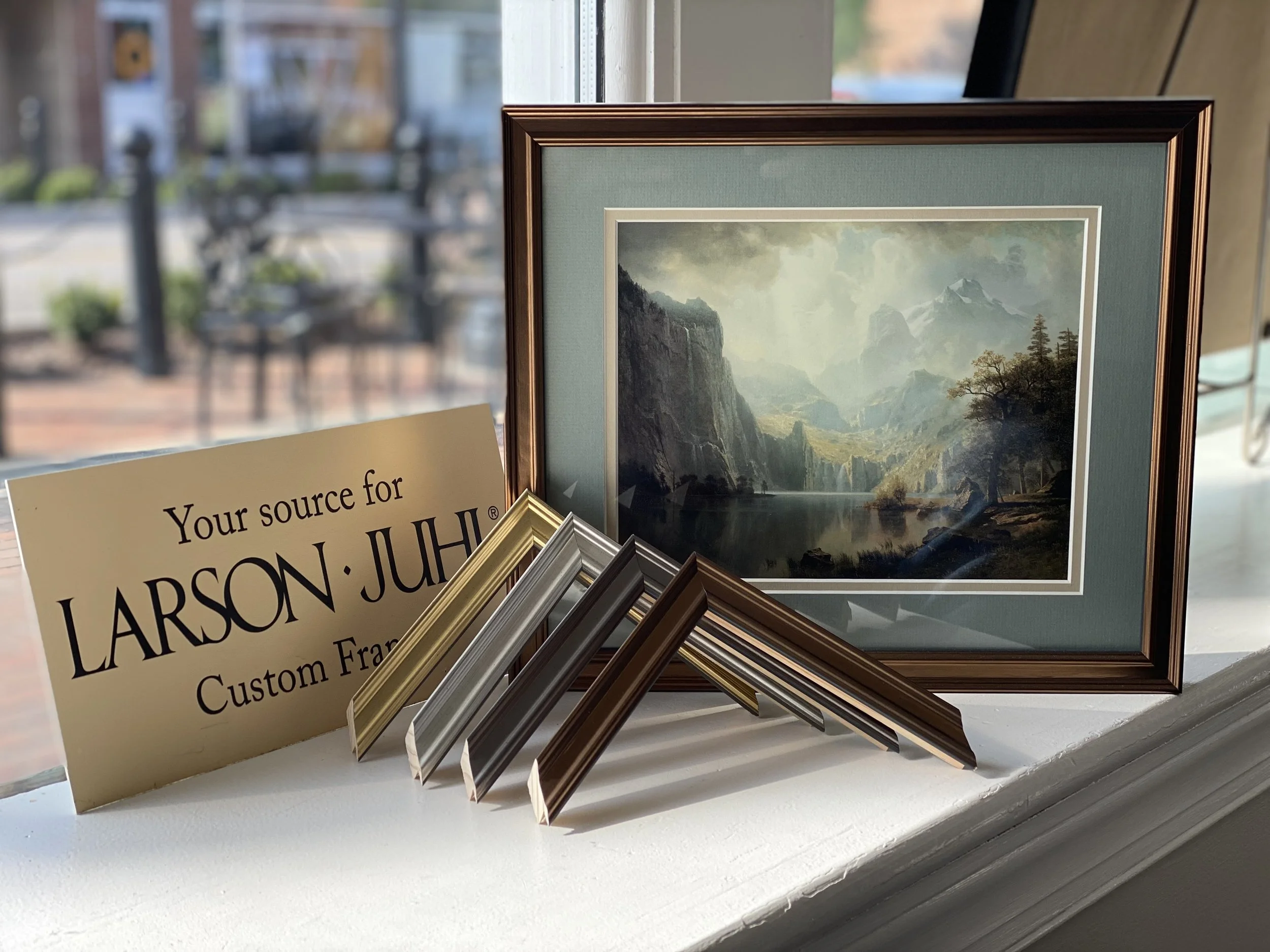 Display of framed landscape paintings and sample picture frames on a white flat surface next to a window.