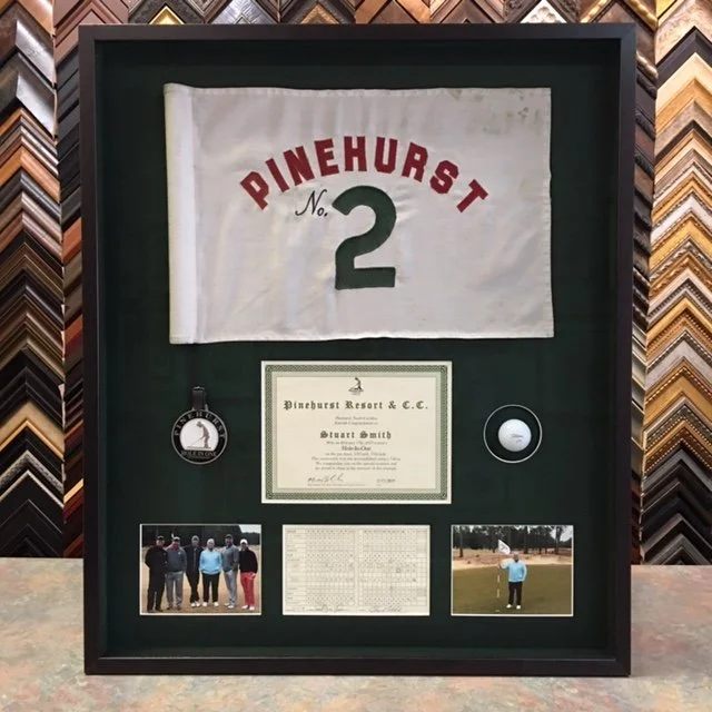 Framed display of a Pinehurst No. 2 golf flag, a certificate from Pinehurst Resort, a golf tee, a group photo, a scoreboard, and a golfer on the course.