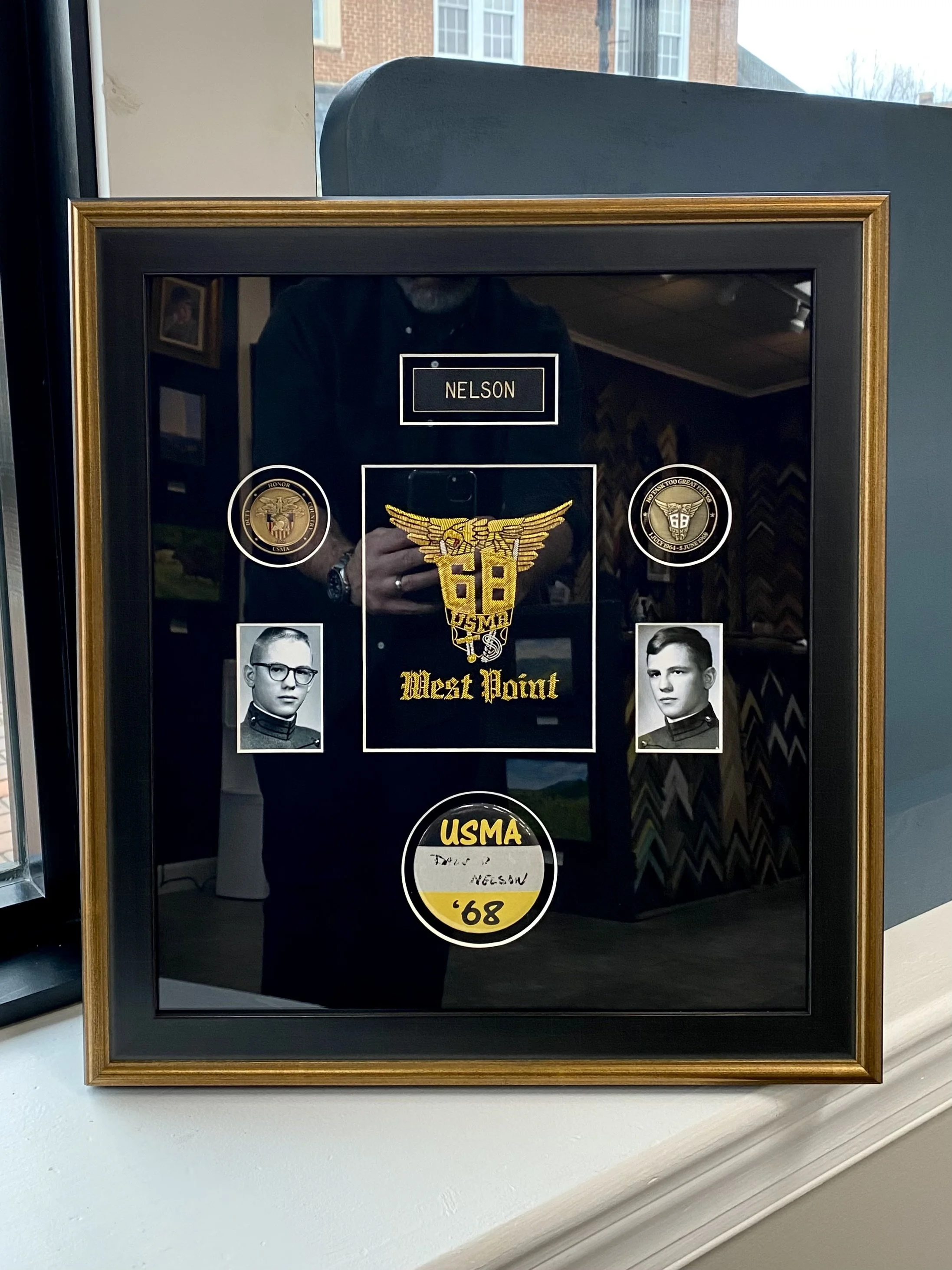 A framed display case with black background and gold trim, showcasing a military insignia, photographs of two soldiers, and various military patches and memorabilia. The insignia reads 'USMA West Point,' and the photographs are black and white portra