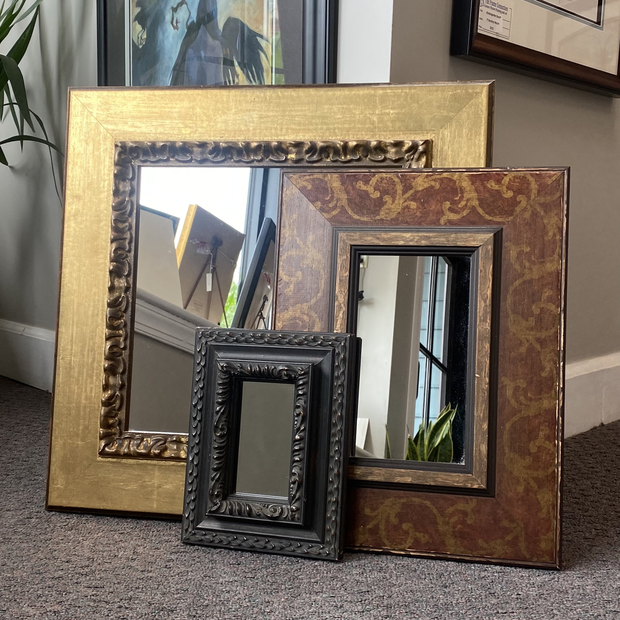 Group of three framed mirrors in gold red and black leaning against a gray wall with dark carpet underneath.