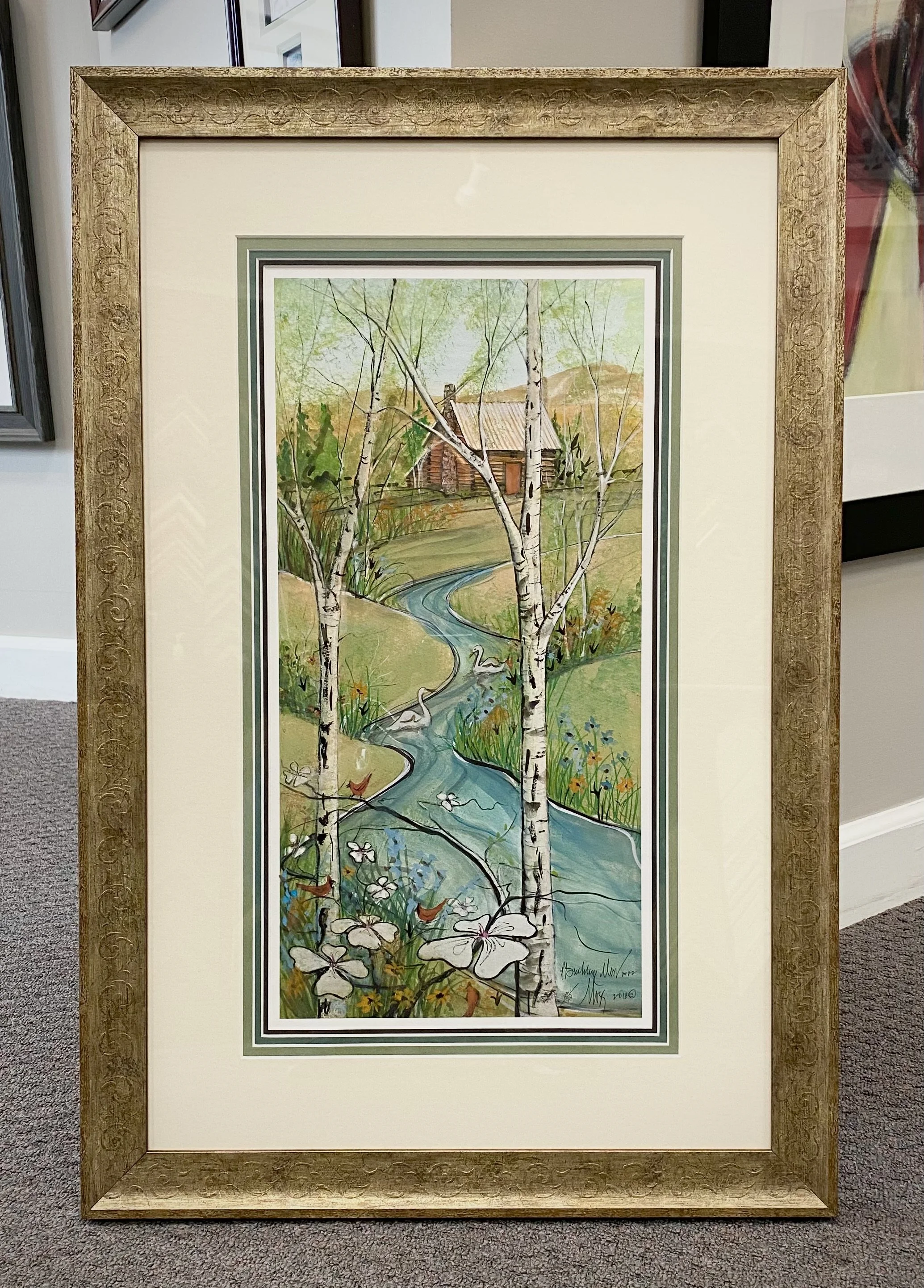 Framed watercolor painting of a rural landscape including a creek, birch trees, wildflowers, a wooden house, and a hill in the background.