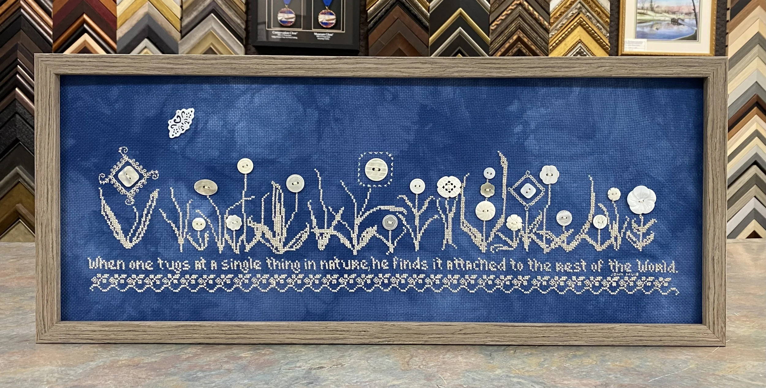Framed artwork with a blue fabric background featuring white embroidery of flowers, grass, and a quote about tugs in nature, with decorative embroidery at the top and bottom.