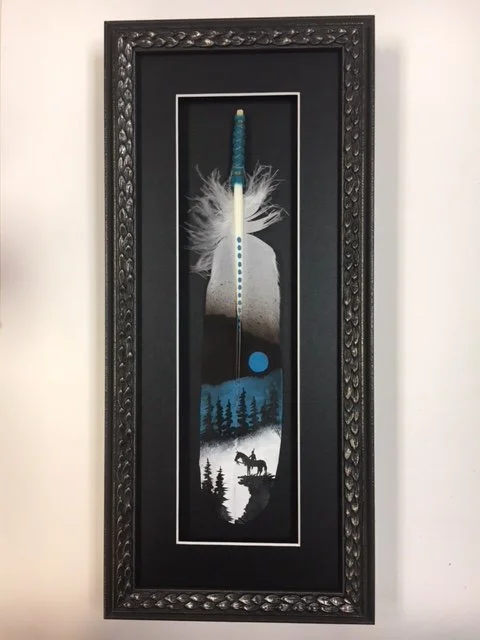 A framed artwork featuring a needle with a feather at the top, a moon and trees, and a dog by a lake, arranged vertically.