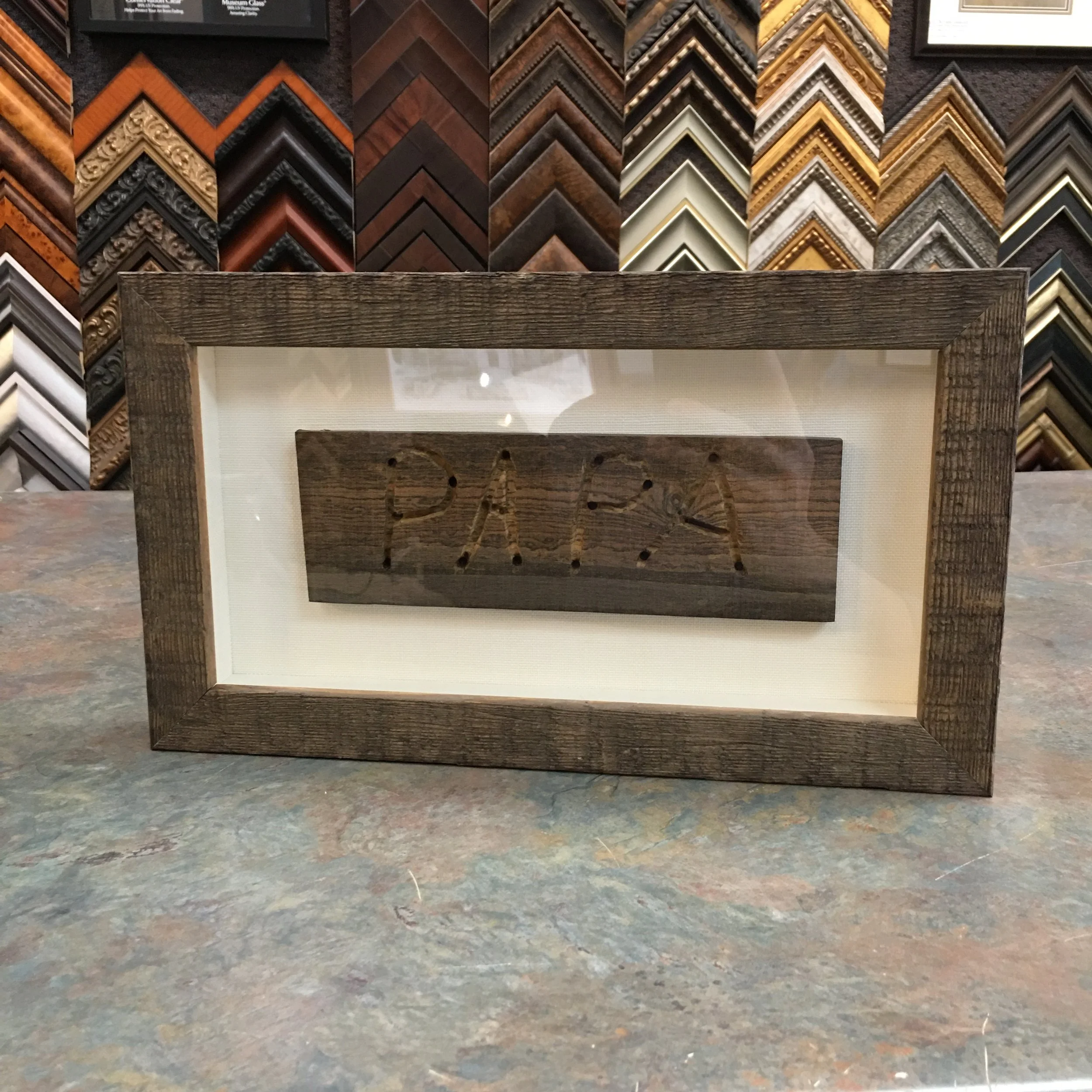 Wooden picture frame with glass, positioned on a colorful speckled countertop, with a display of picture frame samples in the background.