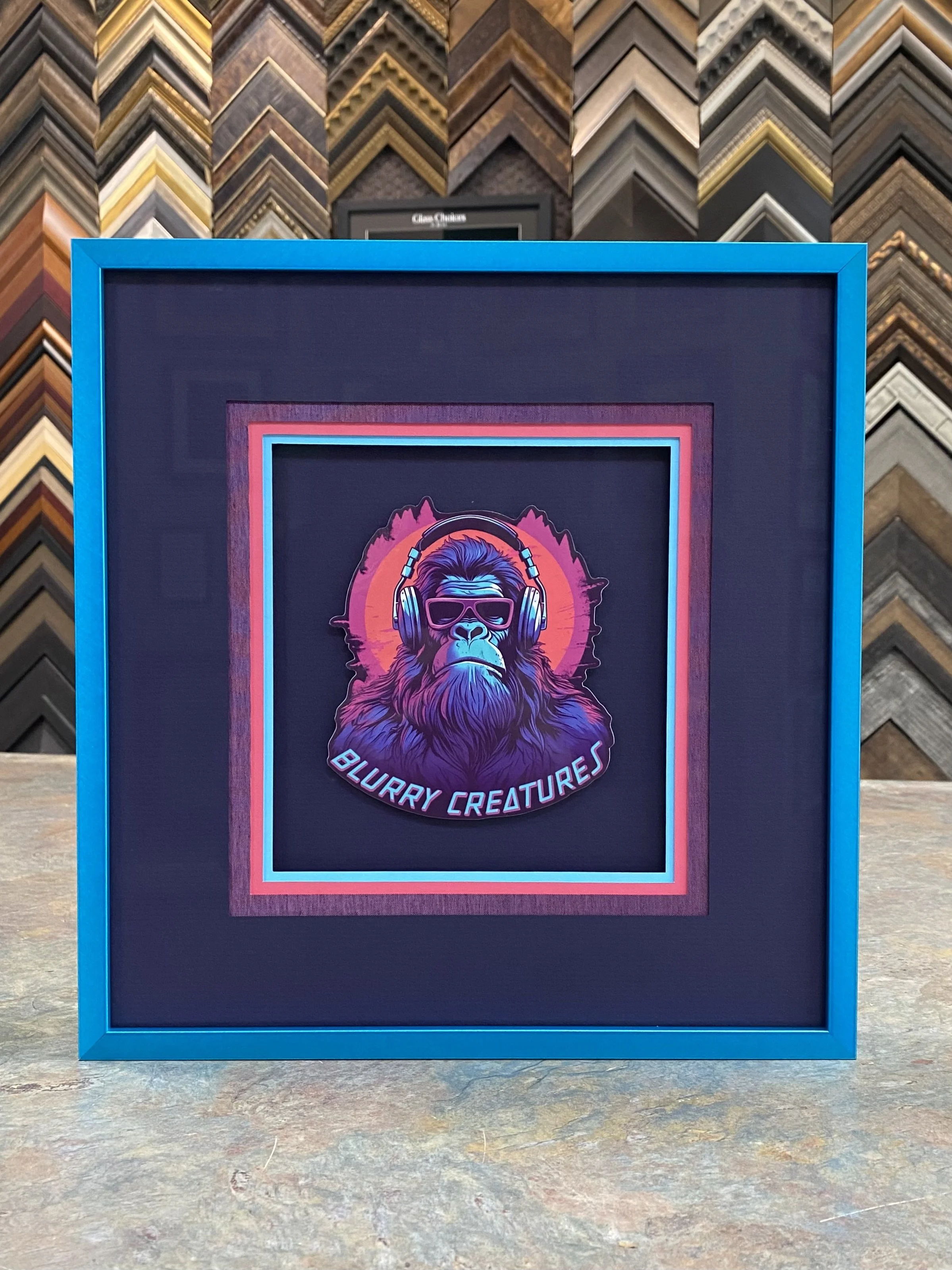 A colorful poster featuring a stylized illustration of a gorilla wearing headphones and sunglasses, with the text 'Blurry Creatures' underneath, framed in blue, set against a background of picture frame samples.