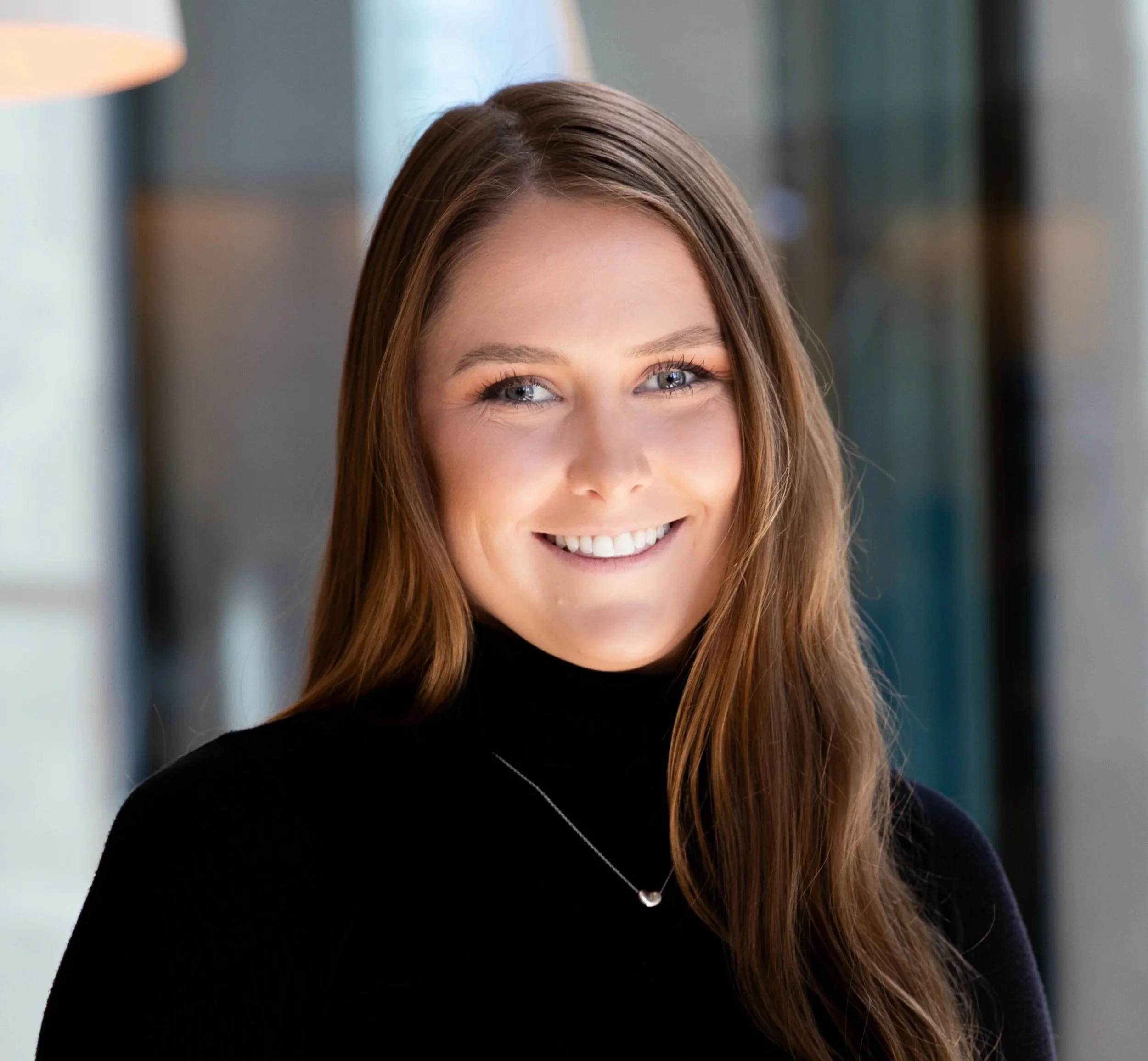 Close-up of a young woman with long brown hair, blue eyes, smiling, wearing a black turtleneck and a silver necklace.