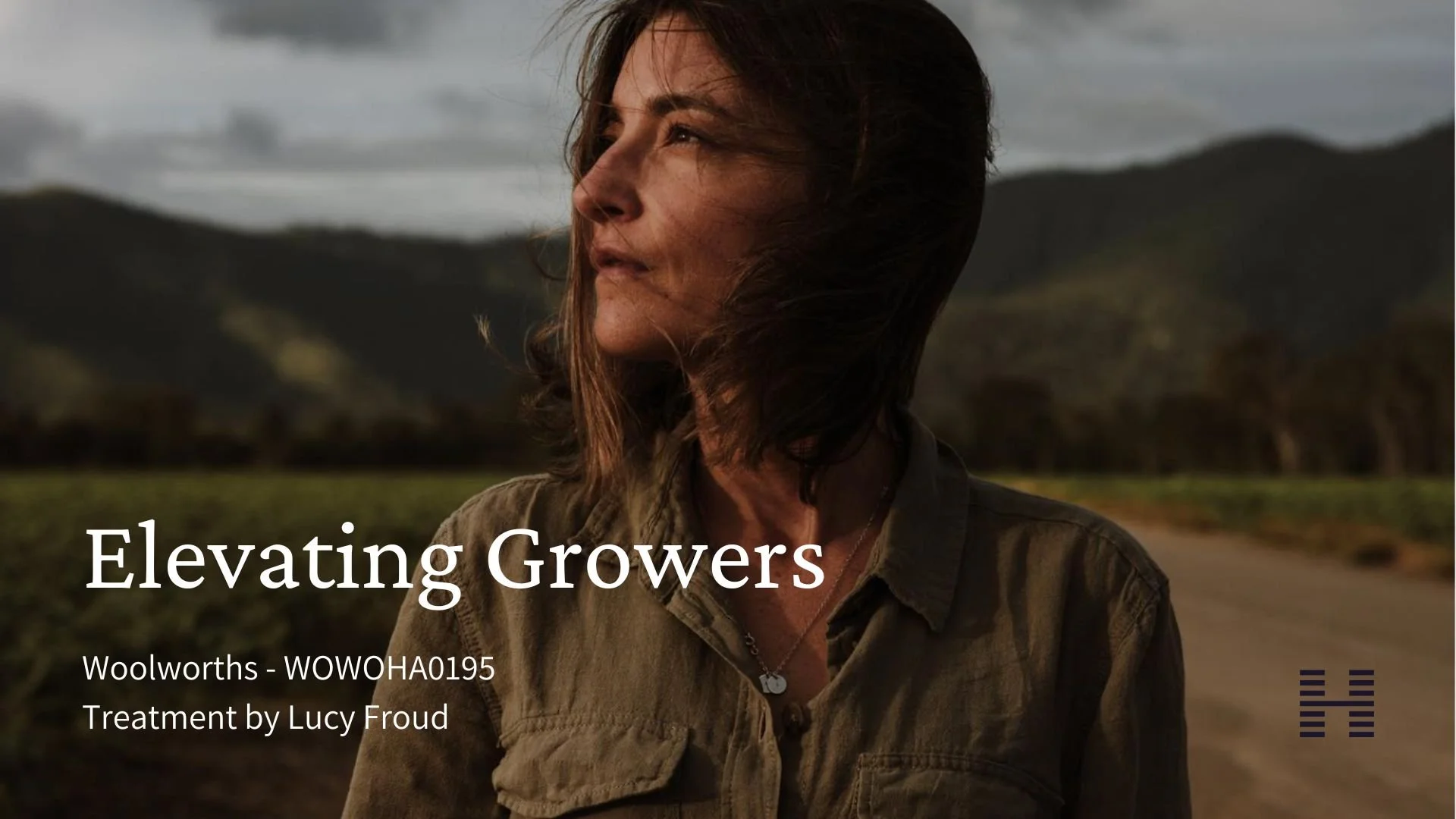 A woman with shoulder-length hair looking into the distance outdoors with mountains and a cloudy sky in the background. She is wearing a tan jacket and a necklace.
