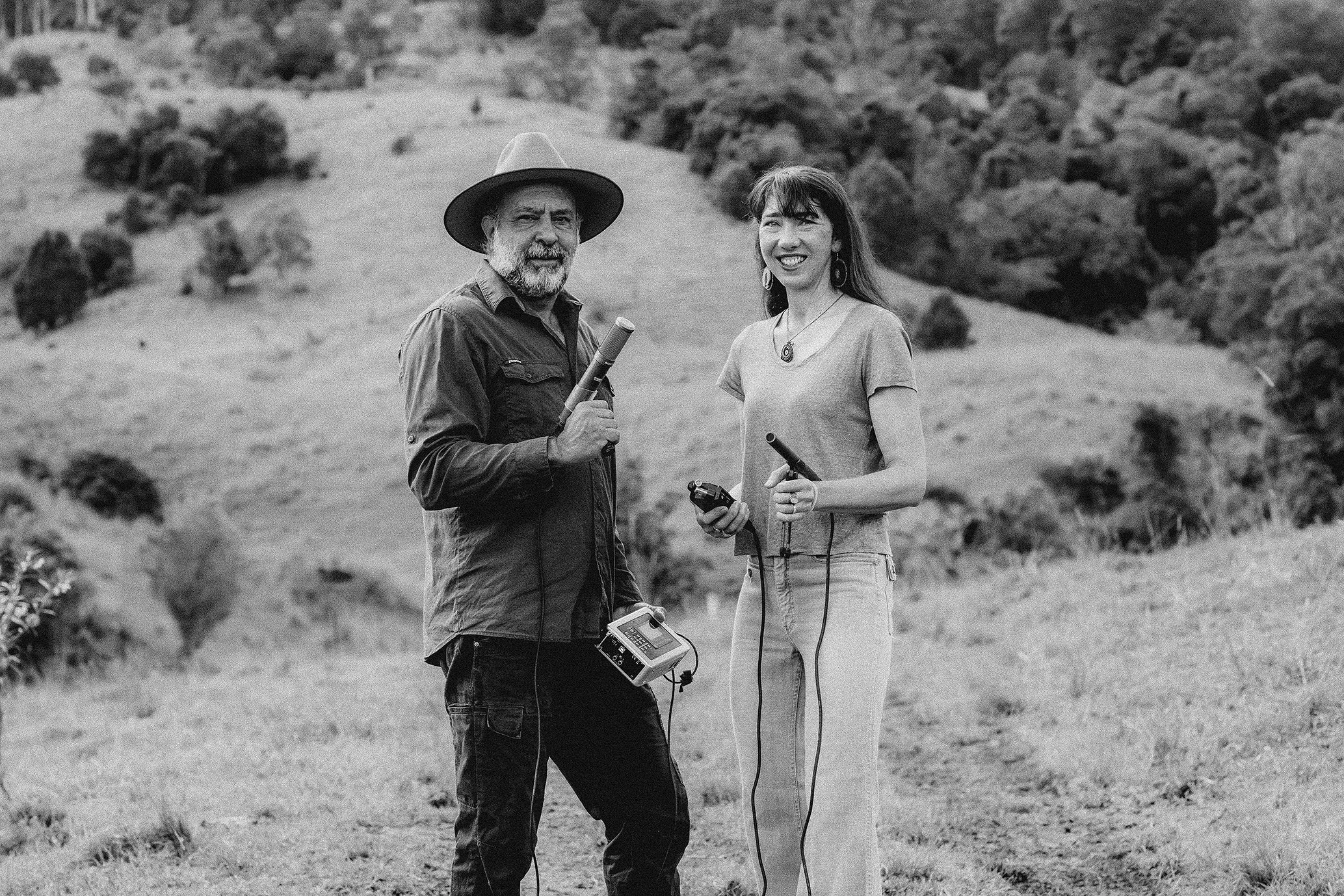 Two people, a man and a woman, standing outdoors in a grassy, hilly area, holding measuring or scientific instruments, smiling at the camera.