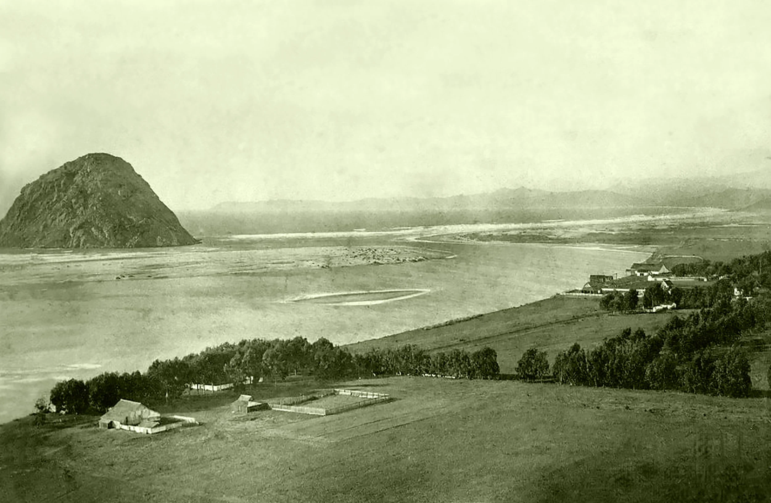 A black and white landscape photo showing a mountain on the left, a large flat floodplain with a meandering river, and a small rural community with farm buildings and trees in the foreground.