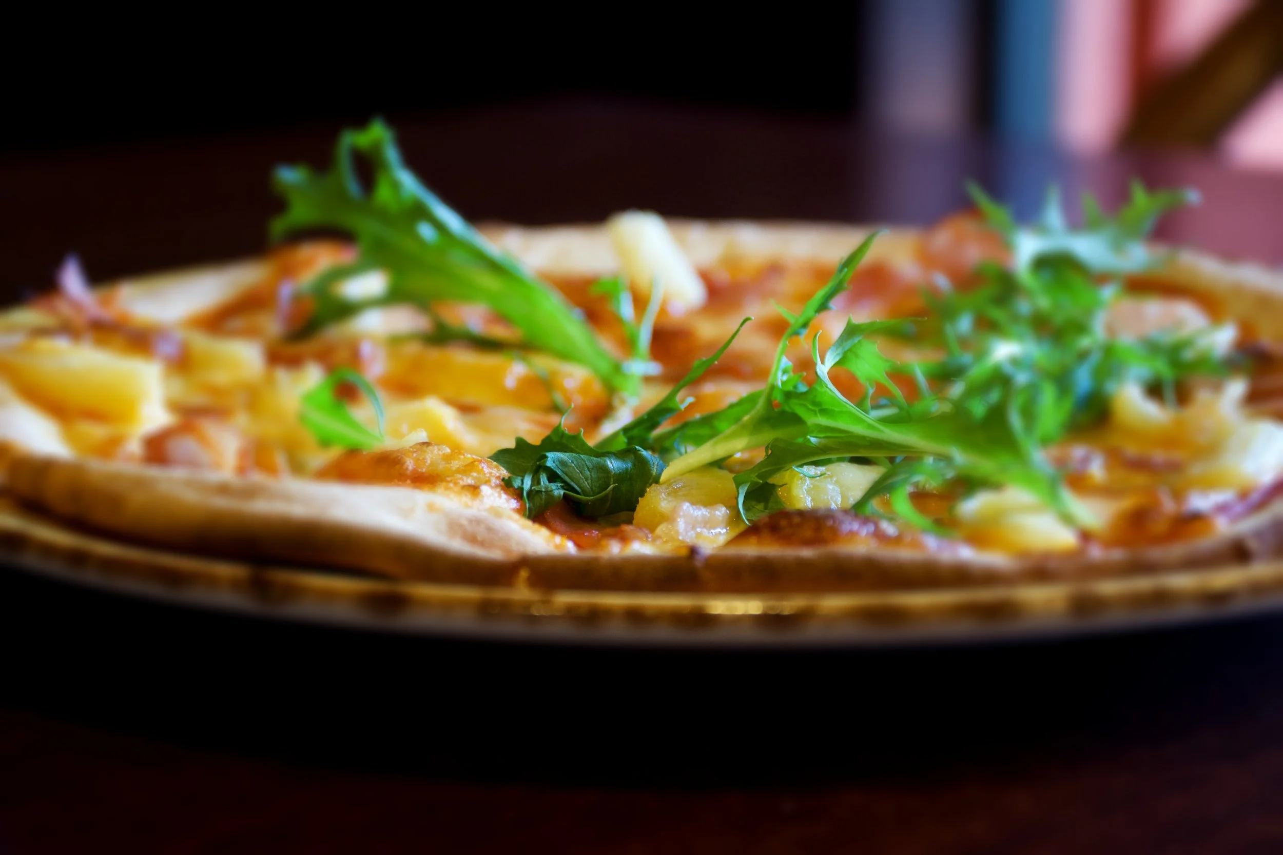 A close-up view of a pizza topped with cheese, bacon, and fresh green arugula leaves on a dark surface.