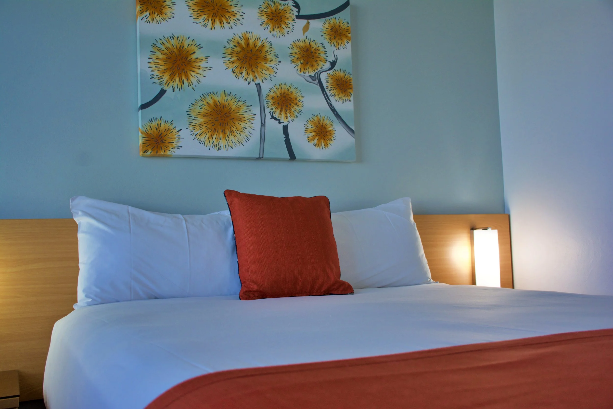 A neatly made hotel bed with white sheets, two white pillows, and a red accent pillow. A wooden headboard and a small bedside lamp are visible, with a colorful floral painting hanging above the bed.