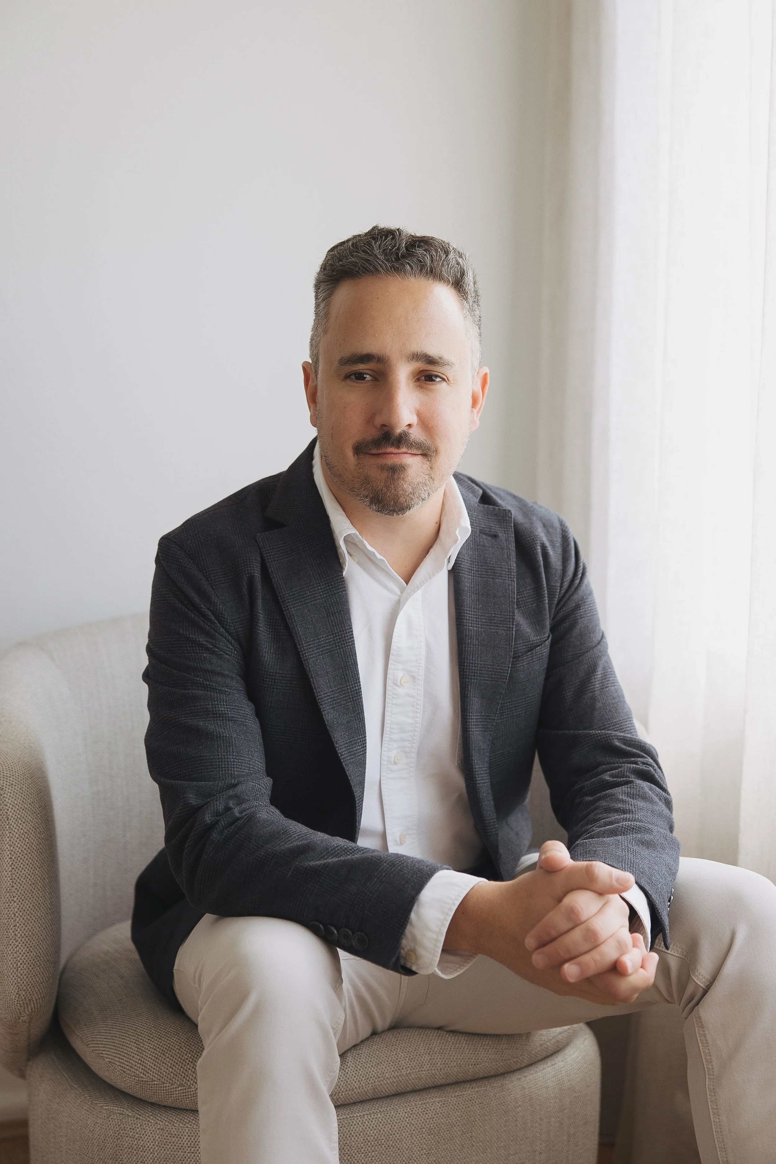 A man seated on a beige armchair with a light-colored background and sheer curtains, wearing a dark blazer over a white shirt and beige pants.