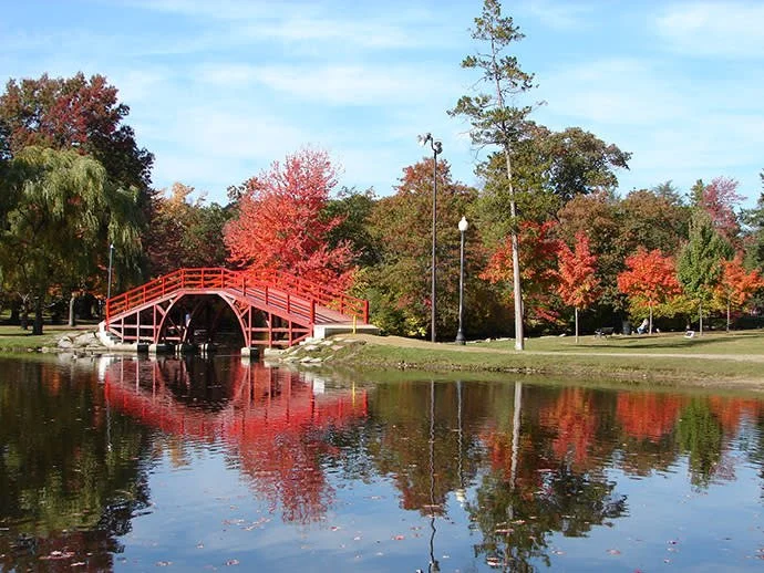 Elm Park, Worcester Mass, Red arched bridge over a pond with colorful autumn trees and a clear blue sky in a park.