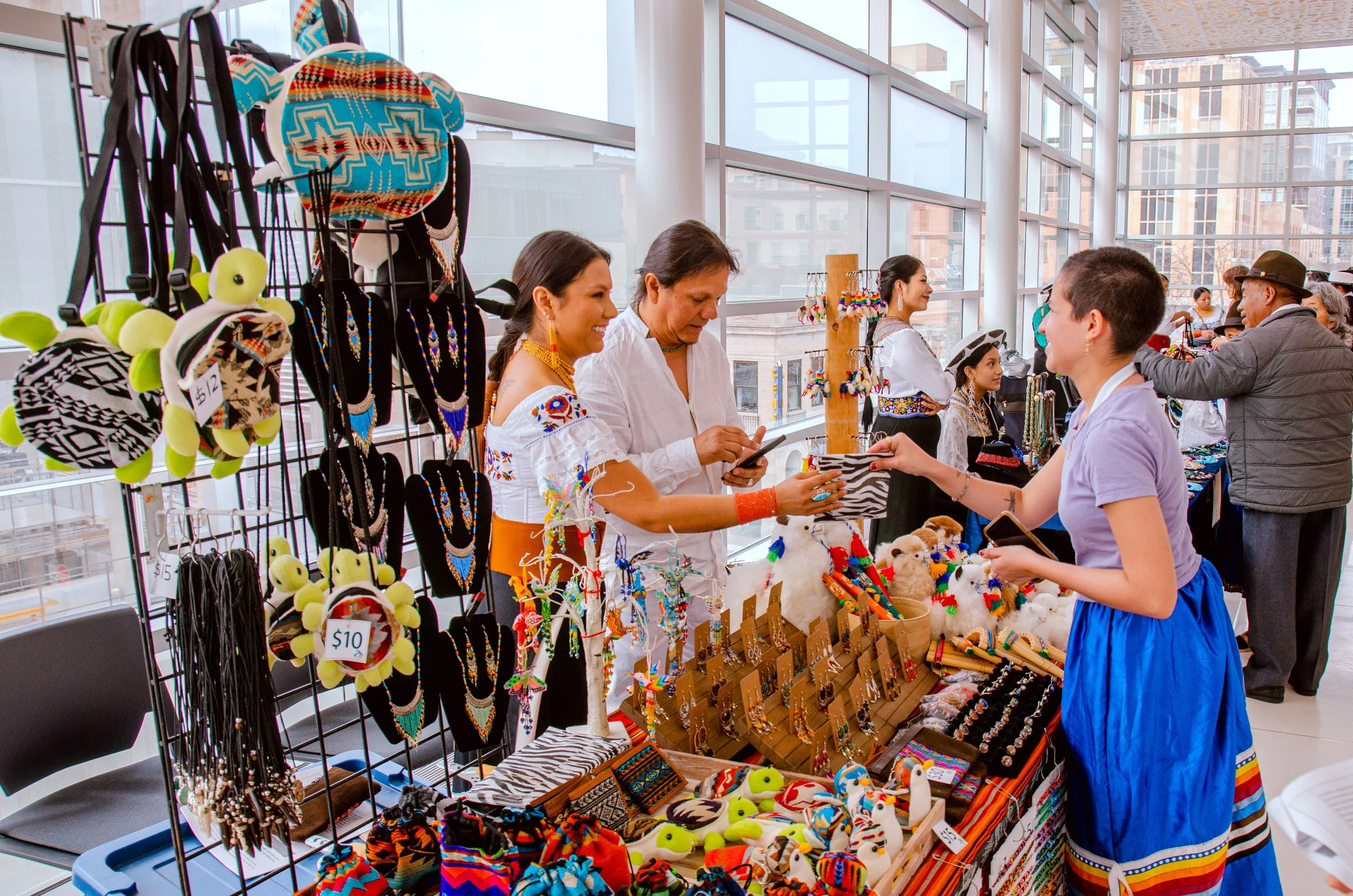 A craft market stall with various handmade jewelry, plush toys, and accessories, featuring vendors dressed in traditional Mexican clothing, and customers browsing inside a modern, glass-walled building.