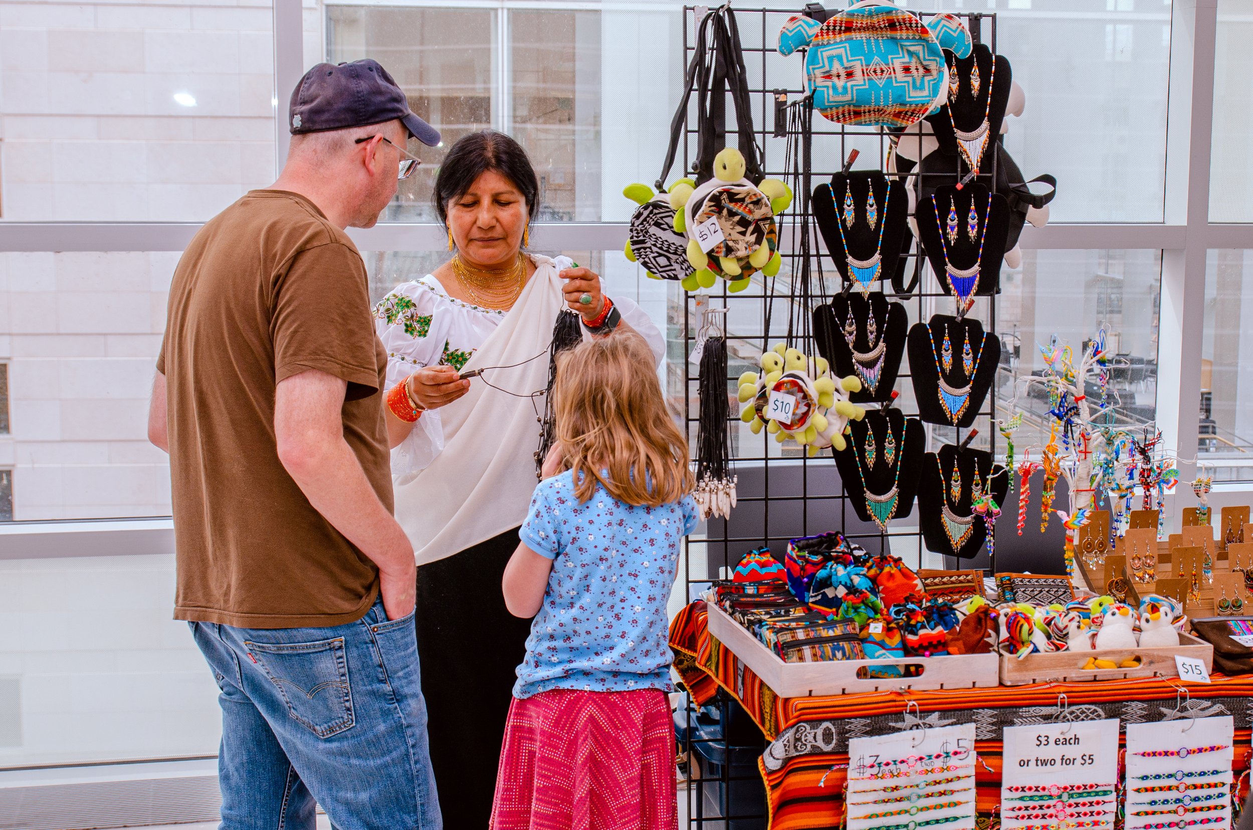 A woman selling handmade jewelry and accessories at a street market stall. She's showing necklaces to a man and a young girl. The stall has various jewelry, hats, and decorative items on display with price tags.
