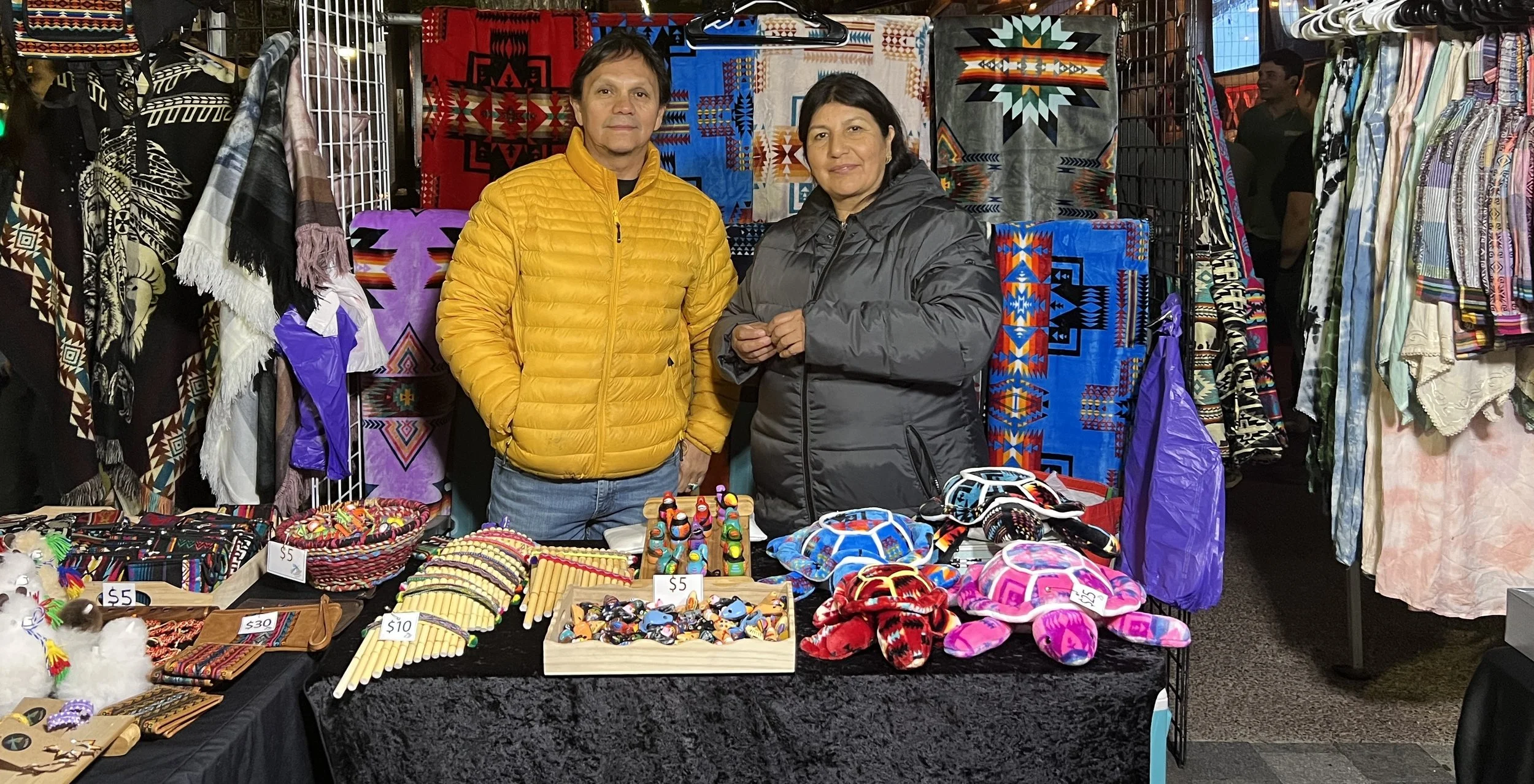 Two people standing behind a table at a market stall selling colorful textiles and handmade crafts. The man is wearing a yellow jacket, and the woman is in a gray jacket. The background features vibrant patterned blankets and woven textiles, with additional clothing and accessories displayed on racks around them.