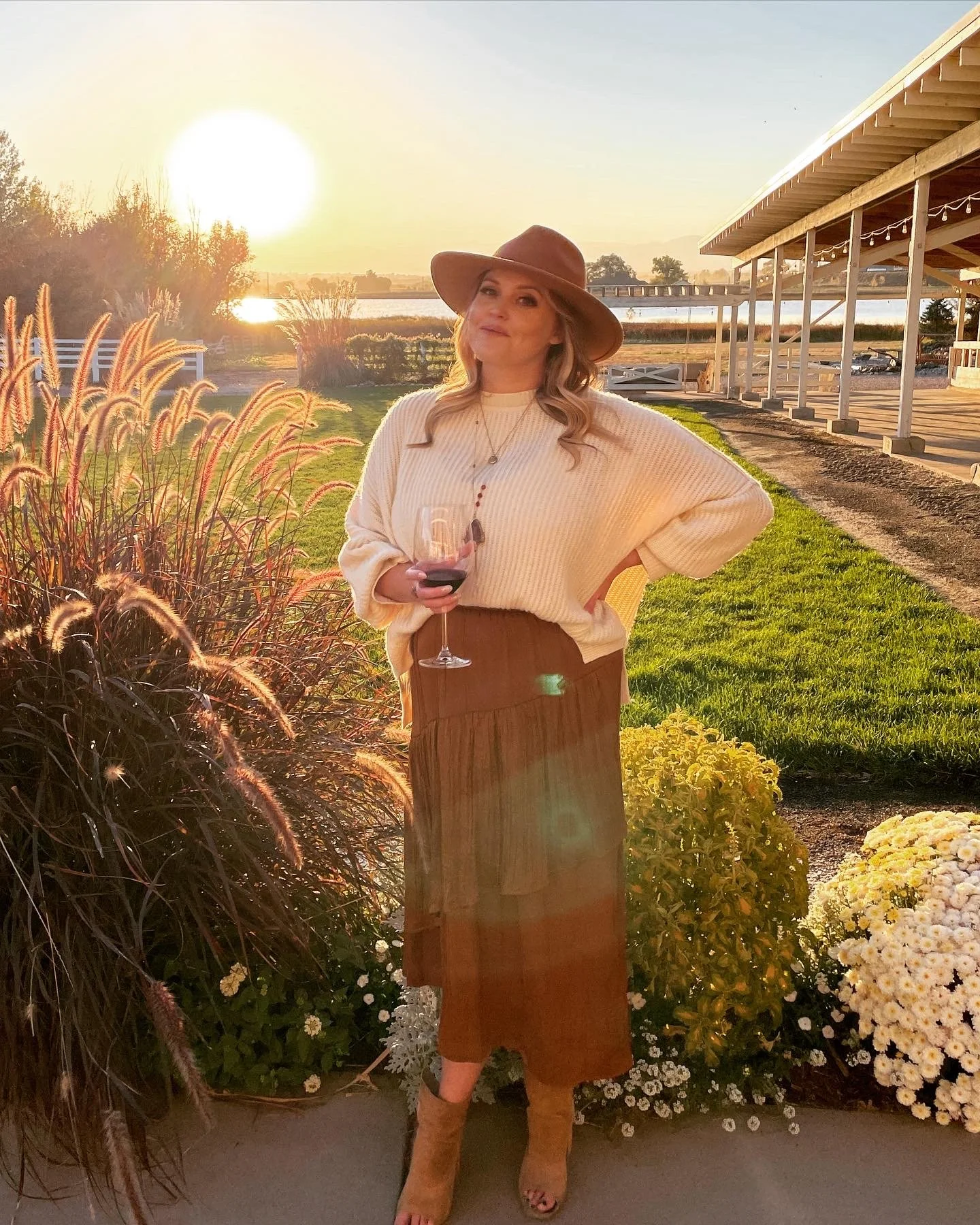 A woman standing outdoors at sunset, wearing a wide-brimmed hat, cream sweater, brown skirt, and boots, holding a glass of red wine with a river and open-air structure in the background.