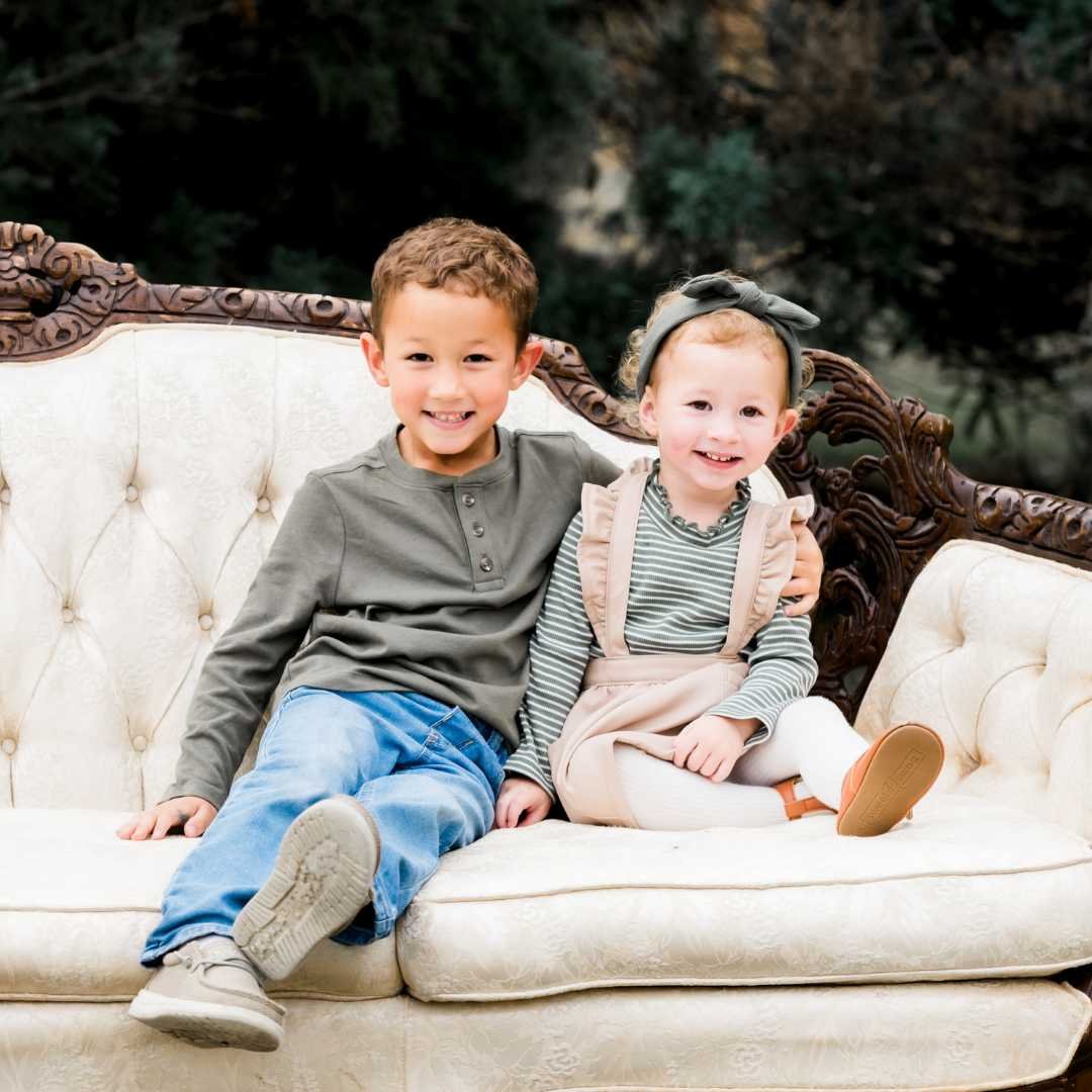 Two smiling children, a boy and a girl, sitting on a vintage cream-colored sofa outdoors with trees in the background.