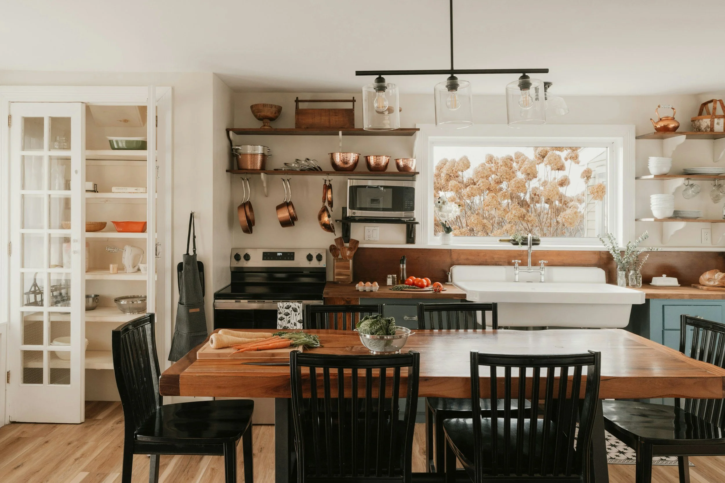 A cozy kitchen with a wooden dining table, black chairs, open shelves holding copper pots, white dishes, and a window with a view of brown trees outside.