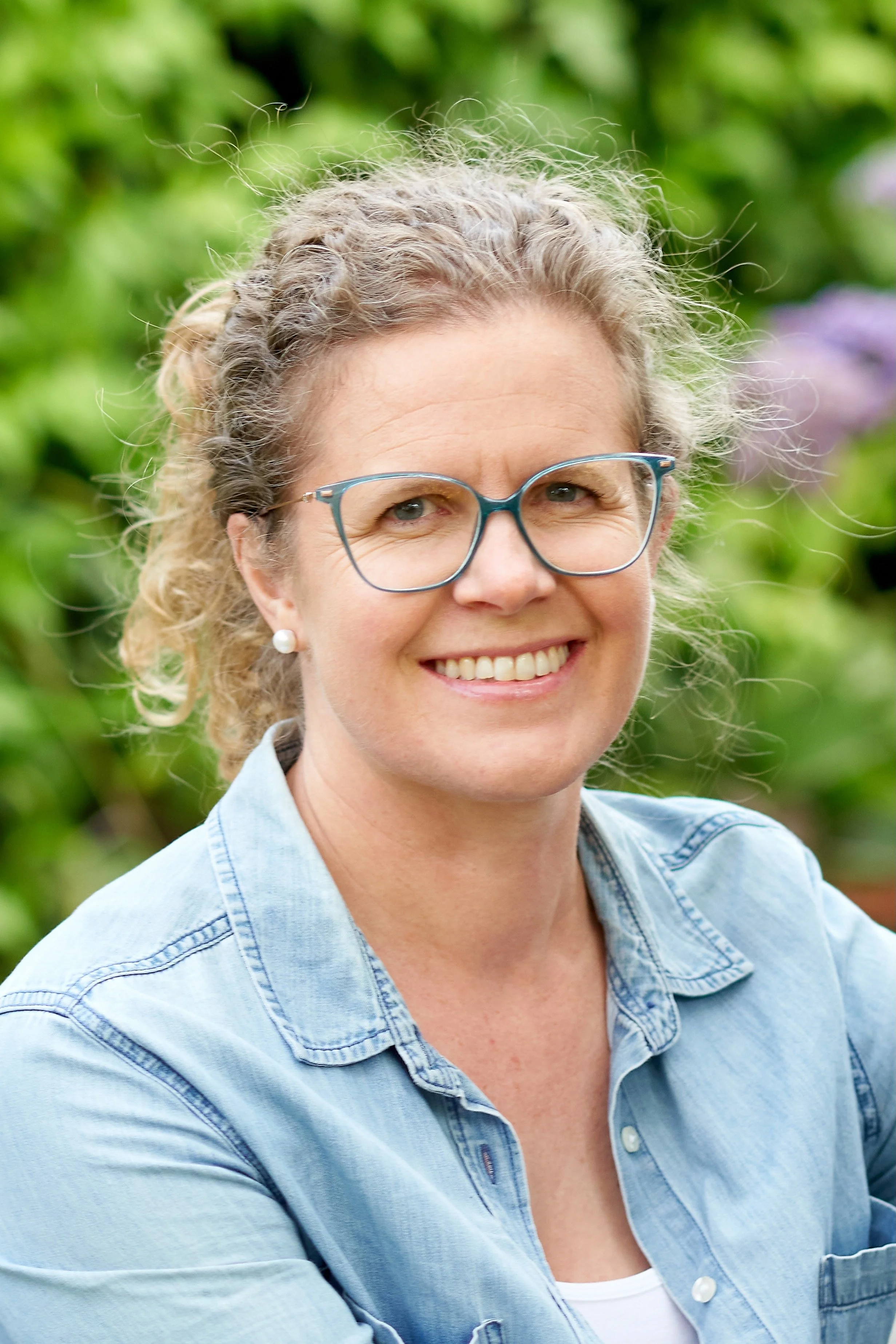 Smiling woman with curly gray hair wearing glasses, pearl earrings, a blue denim shirt, and a white top, outdoors with green foliage in the background.