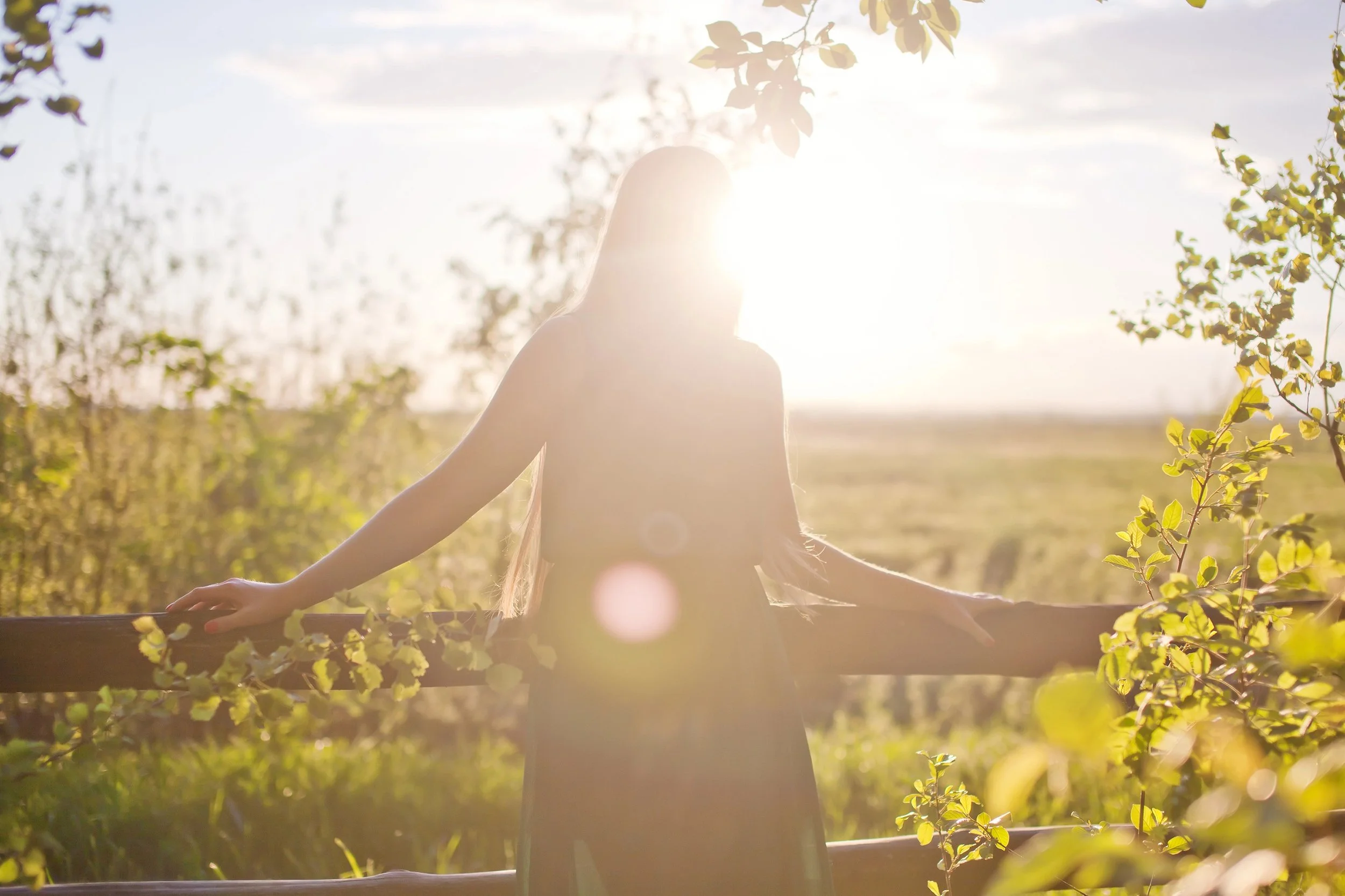 A woman standing outdoors near a wooden fence with arms outstretched, facing the sun at sunset or sunrise, surrounded by trees and foliage.