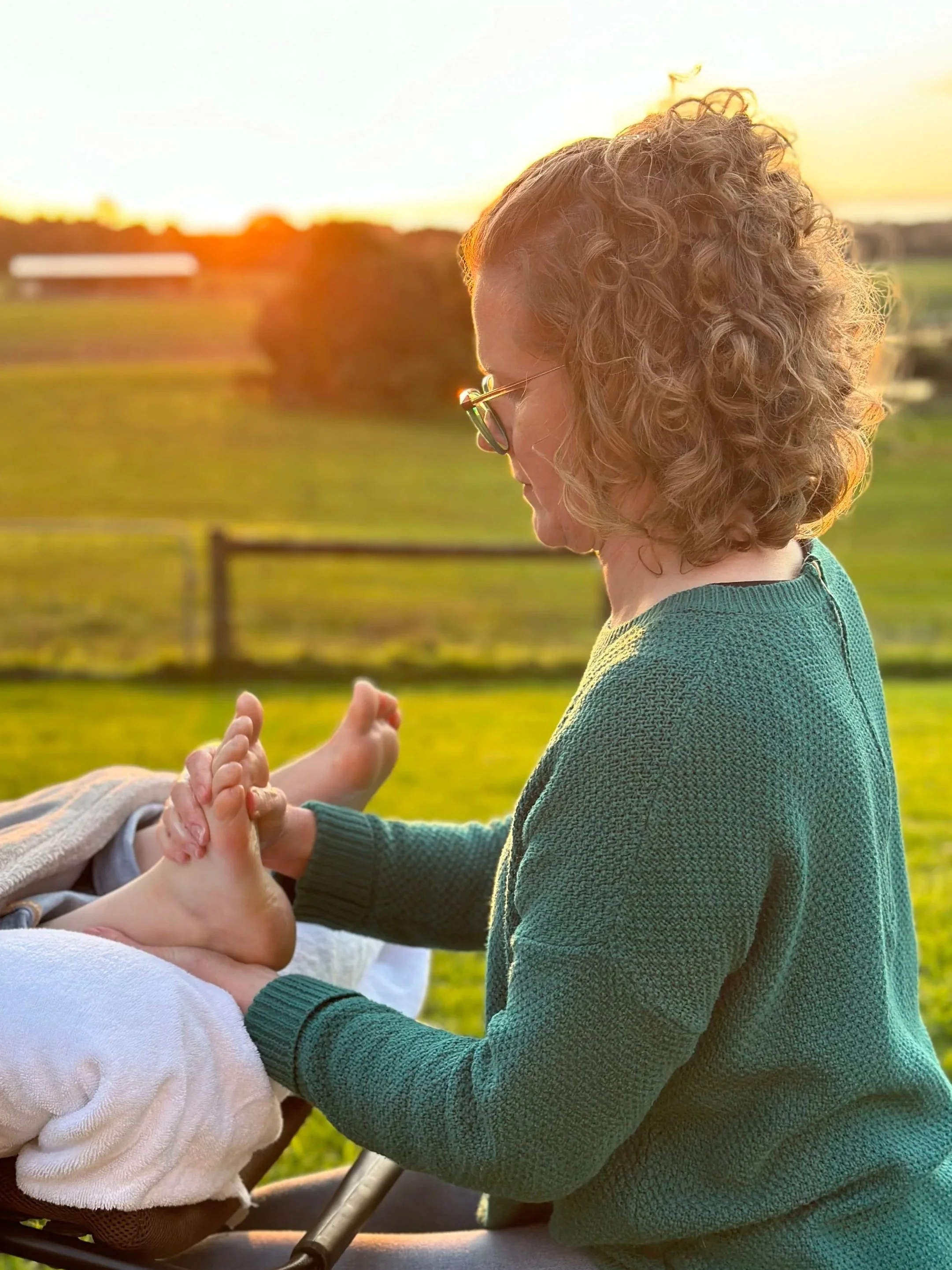 Woman with curly hair and glasses holding the foot of a baby outdoors at sunset