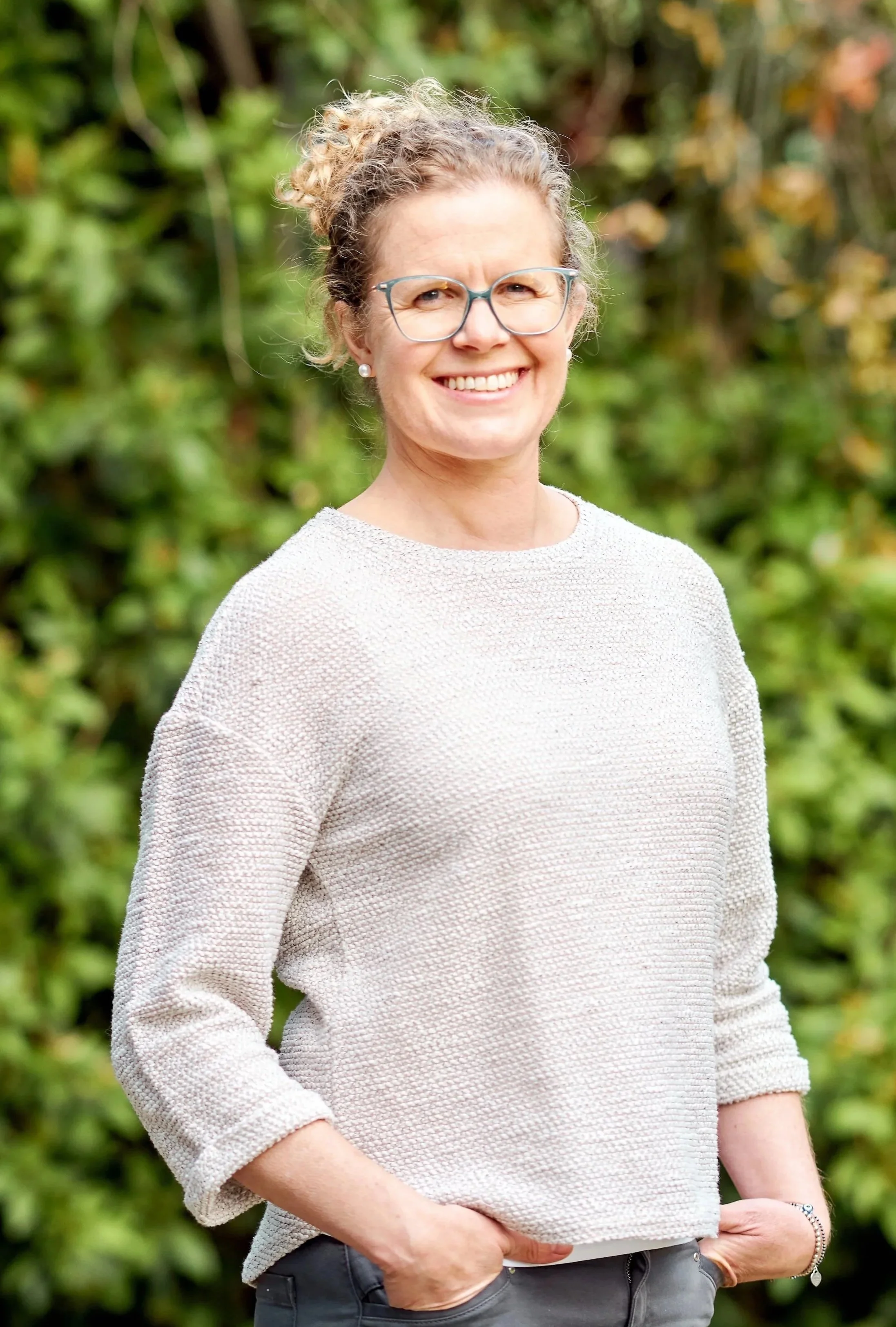A smiling woman with glasses and curly hair tied up, standing outdoors with green foliage in the background.