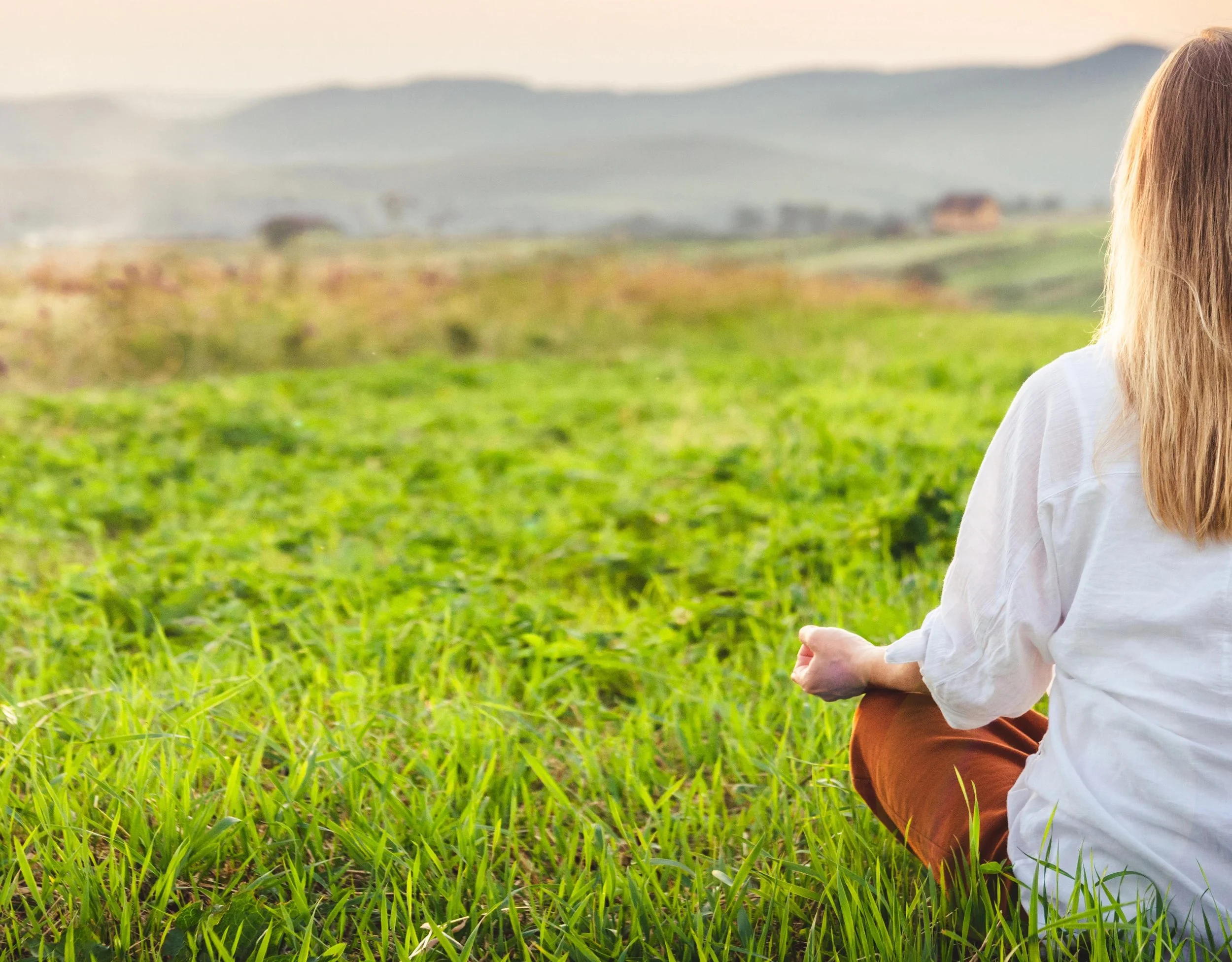 A calm woman with blonde hair sitting cross-legged meditating on green grass in a field during sunset, facing away from the camera with mountains in the background.
