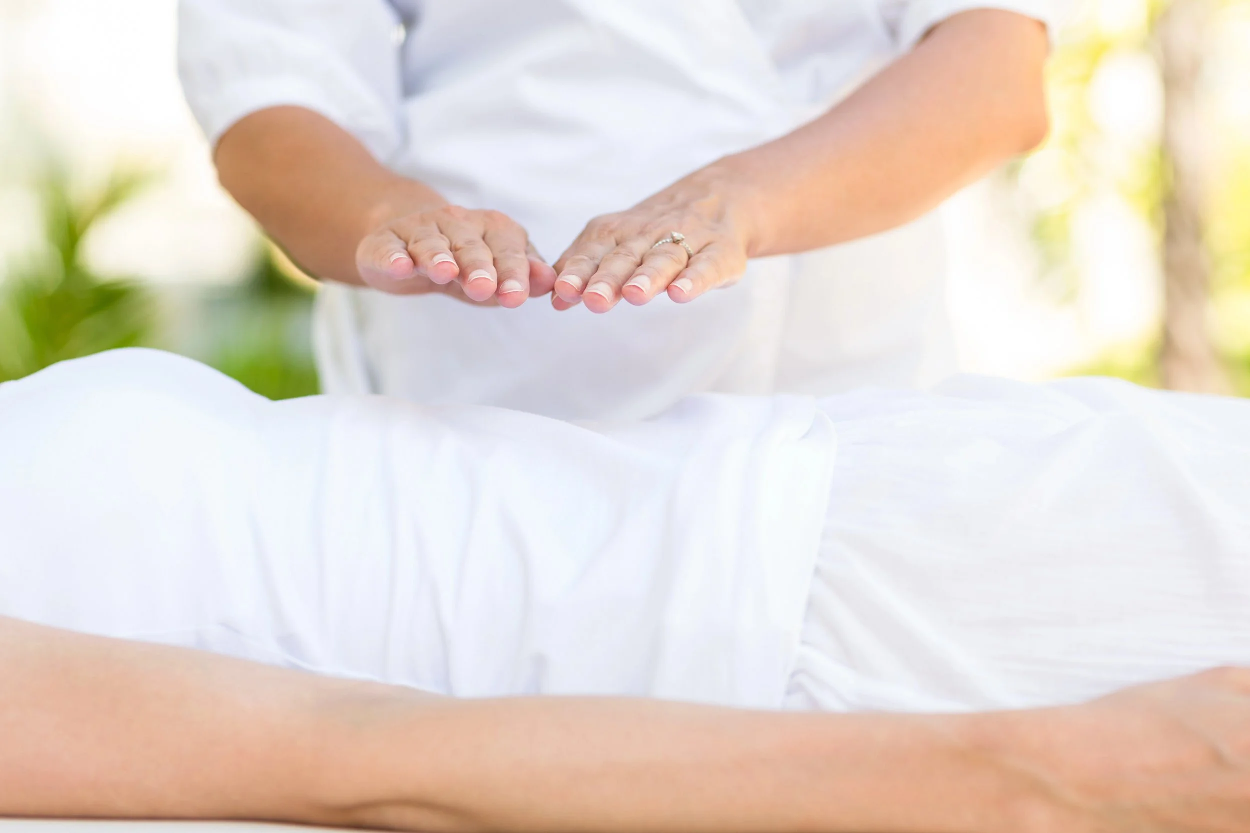Person giving a reiki healing session outdoors, with focus on hands and torso, wearing white clothing, in a natural setting with sunlight and greenery.