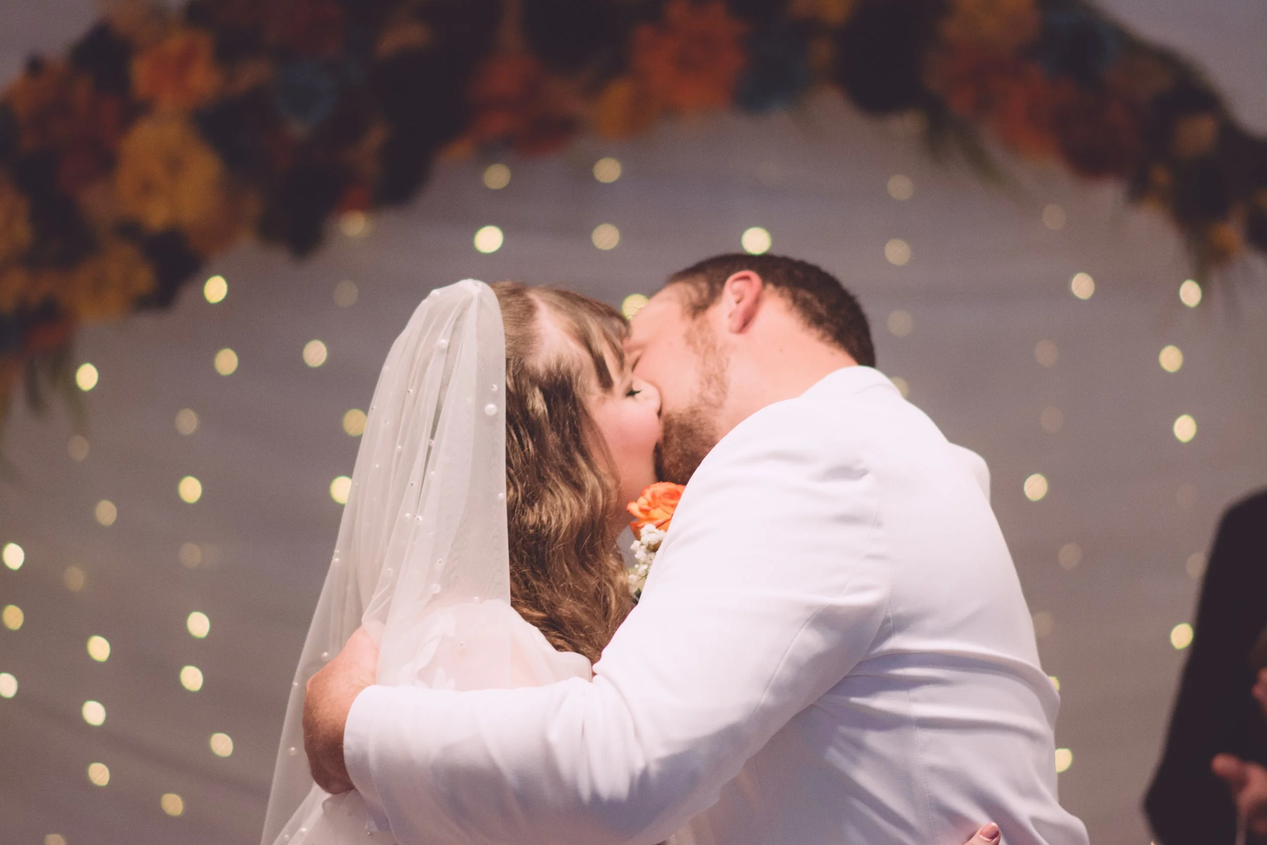 A bride and groom sharing a kiss at their wedding reception under a floral and fairy light decorated ceiling.