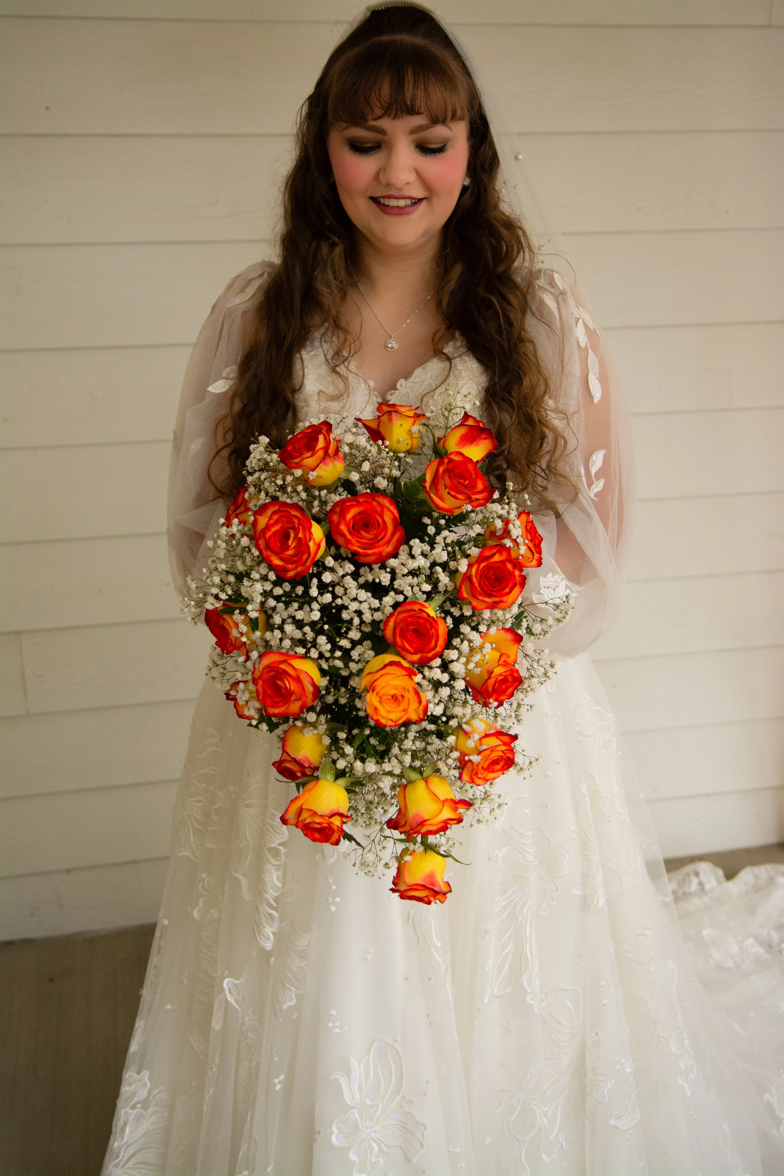 A bride in a white wedding dress holding a cascading bouquet of orange and yellow roses, with baby's breath, standing in front of a white wood-paneled wall.