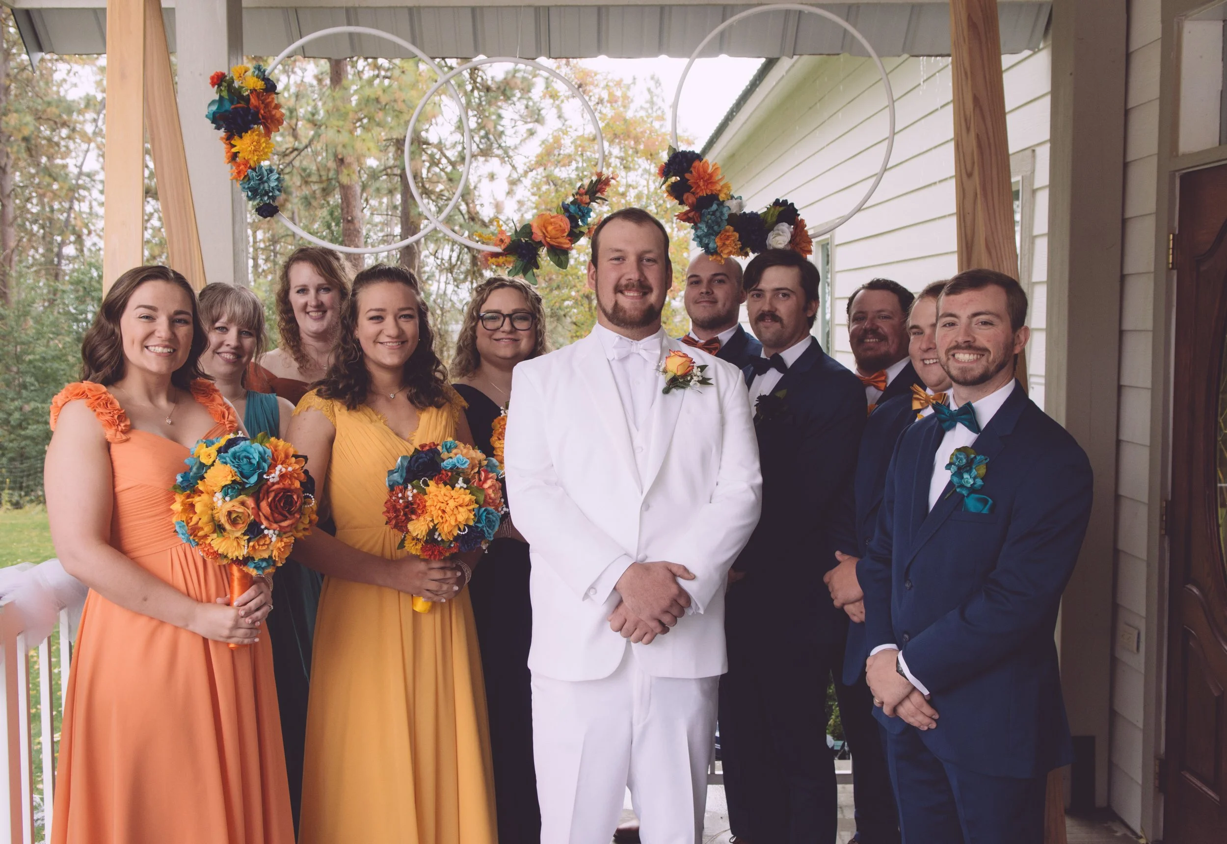 Group of wedding attendees, including bride and groom, posing for a photo on a porch. The bride and many of the bridesmaids are wearing colorful dresses, and the groom and groomsmen are in suits with bow ties. Decorated with floral arrangements and c