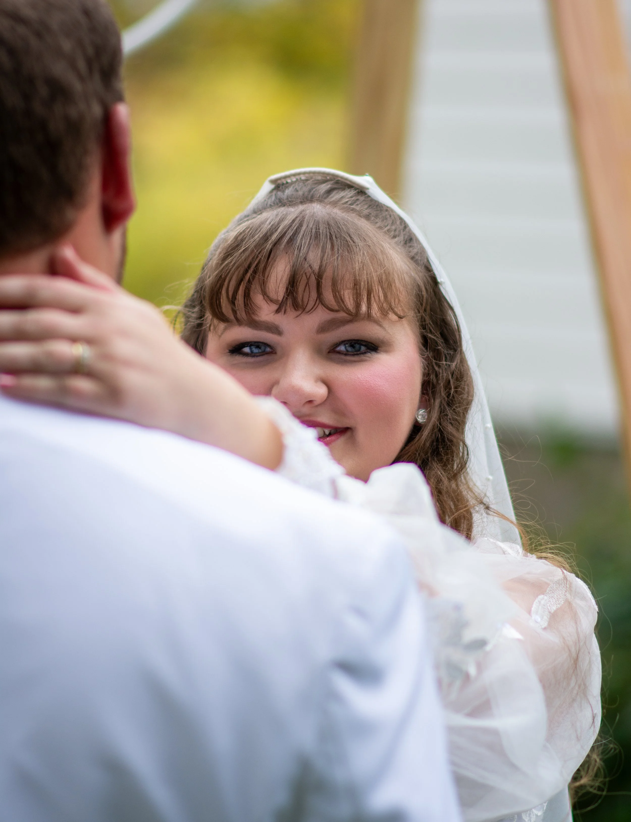 A woman with light brown hair, blue eyes, and a white veil is looking at a man. The woman is smiling gently, and the man, seen from behind, has short brown hair and is wearing a white shirt. The woman's hand is resting on the man's shoulder.