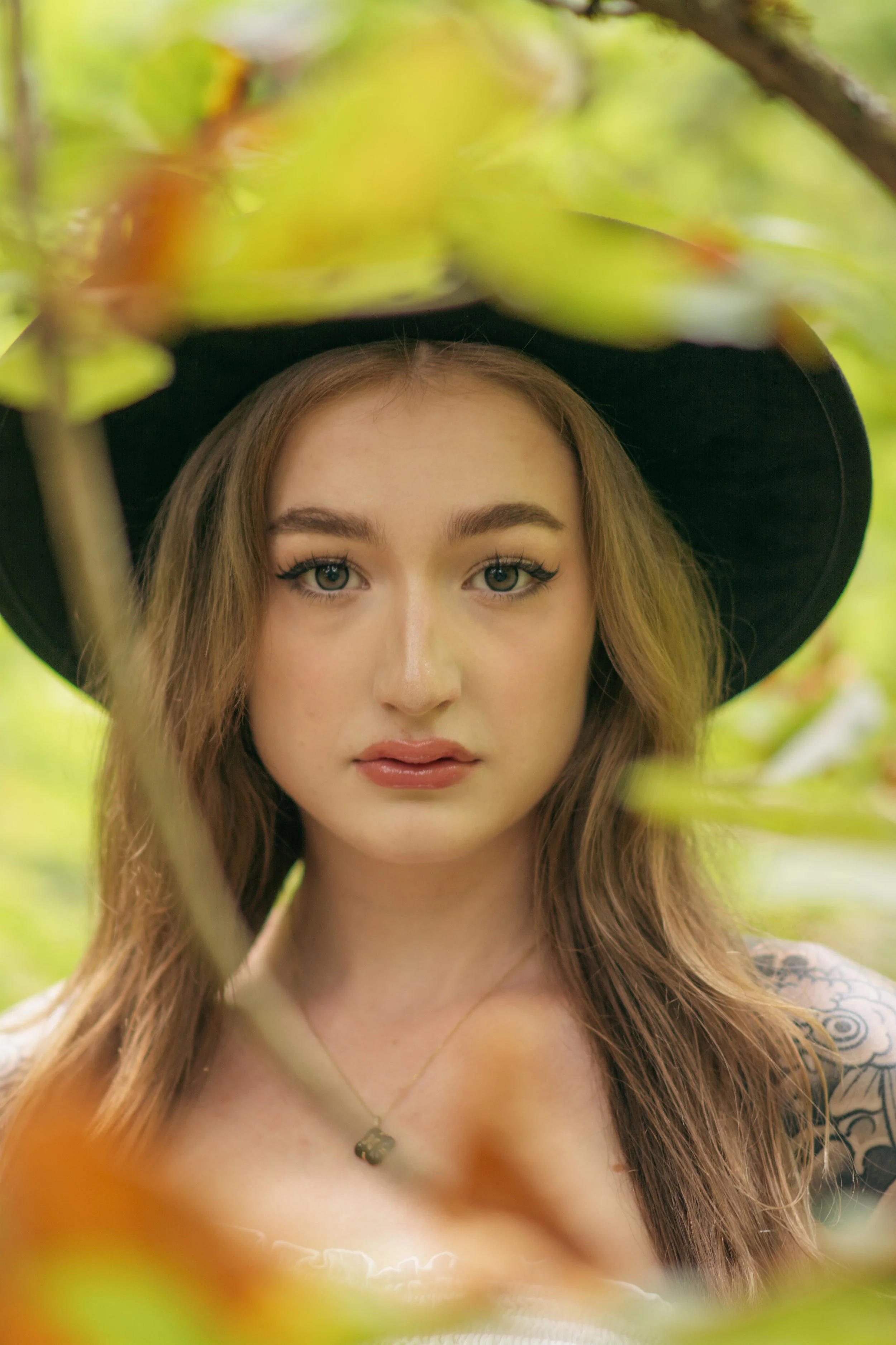 A woman with long brown hair wearing a black hat and a necklace, looking directly at the camera, surrounded by green foliage.