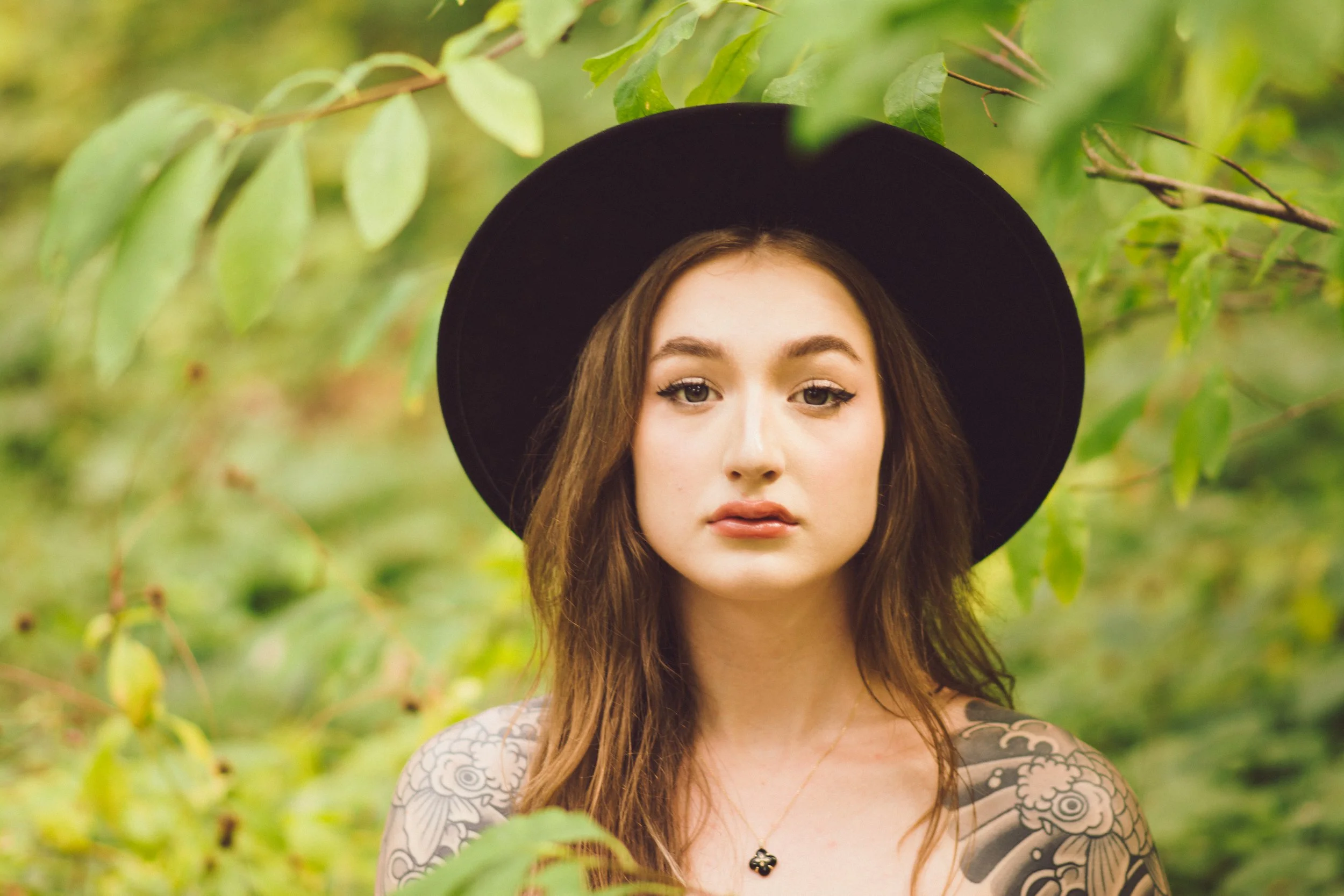 Young woman with long brown hair, wearing a black hat and a necklace, standing among green leaves, some tattoos visible on her shoulder.