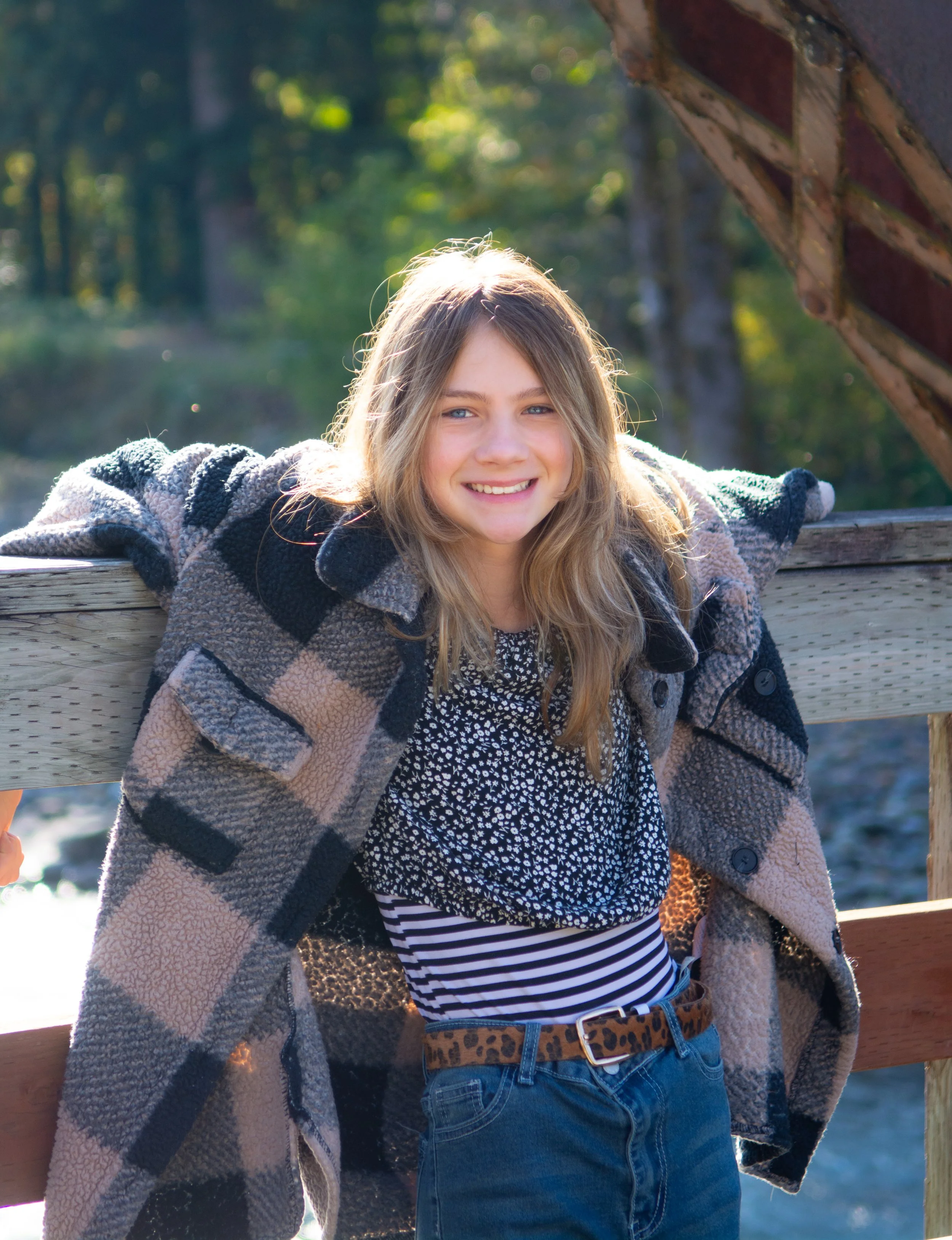 A young girl with long blonde hair, smiling and leaning on a wooden railing outdoors surrounded by trees.
